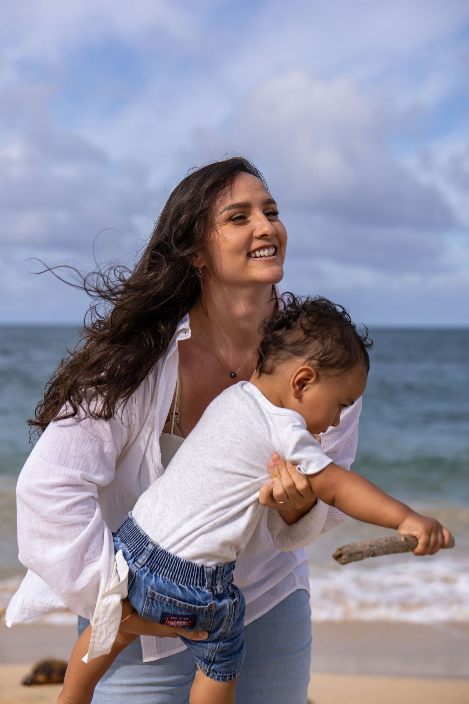 Happy mother holding toddler on a sunny beach with ocean waves behind.
