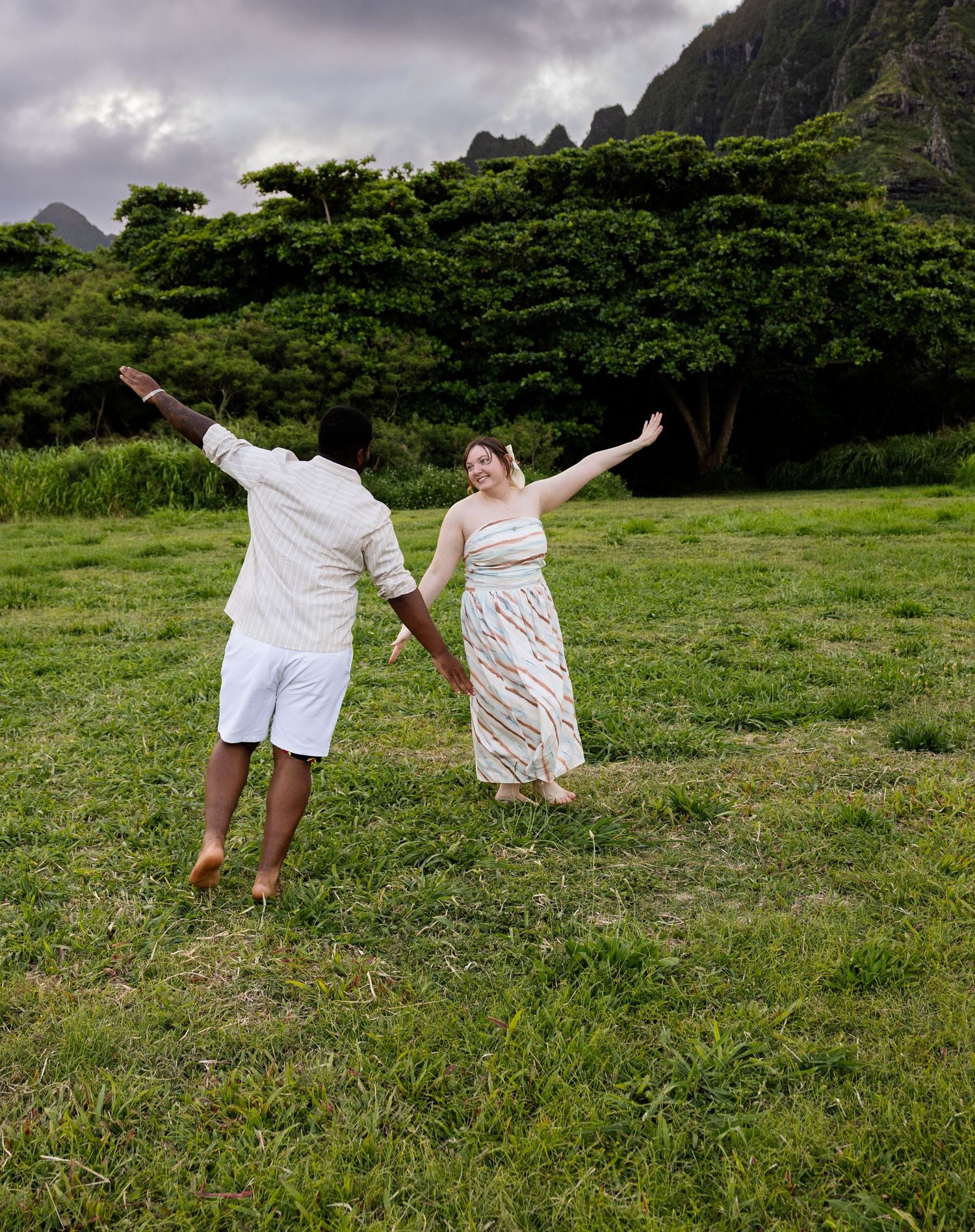 Joyful couple holding hands with arms outstretched in a grassy field with lush Hawaiian mountains.