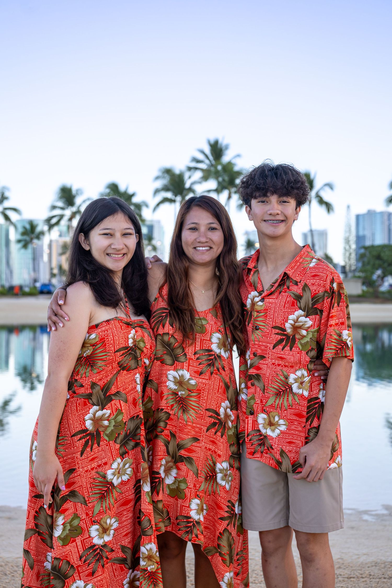 Three people in matching red Hawaiian floral outfits posing near a tropical pool with palm trees and city skyline.