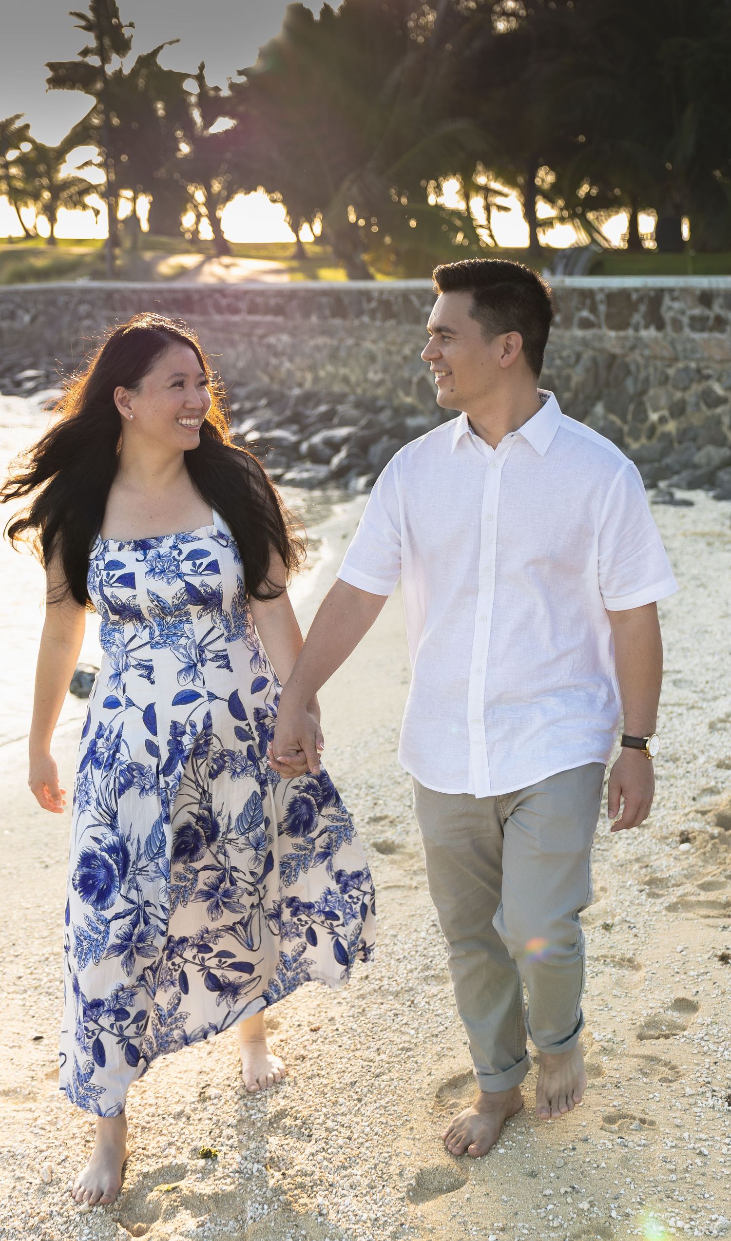 Couple walking barefoot on sandy beach at golden sunset, woman in blue floral dress holding hands.