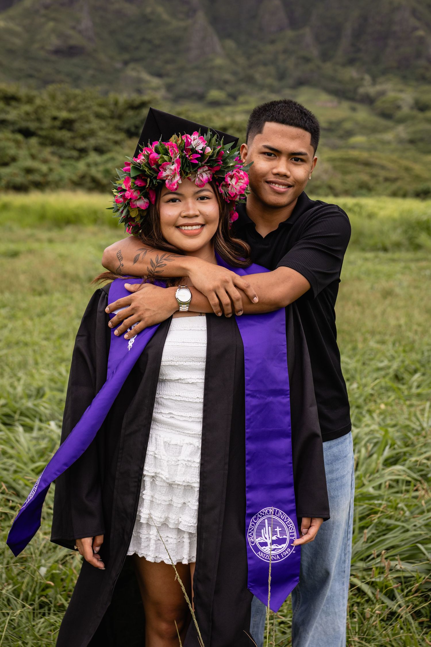 Graduate in black gown and floral lei cap embraced by partner outdoors in Hawaii with mountains behind.