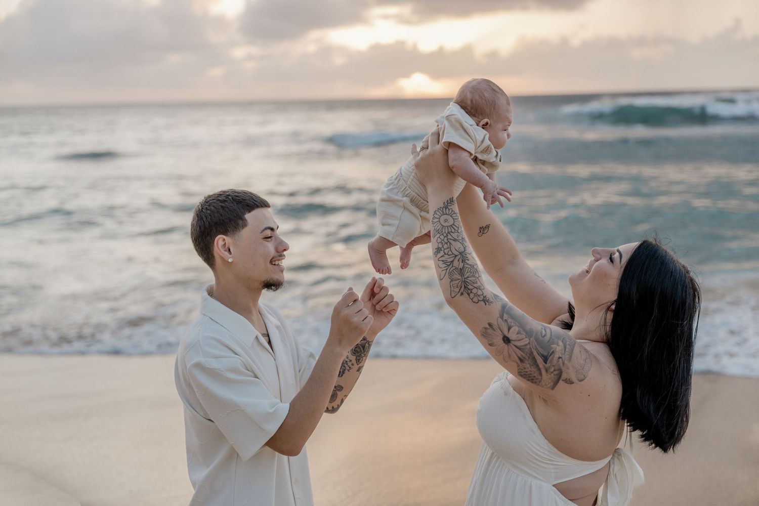 Young family with tattoos lifting baby on beach at sunset, ocean waves in background.