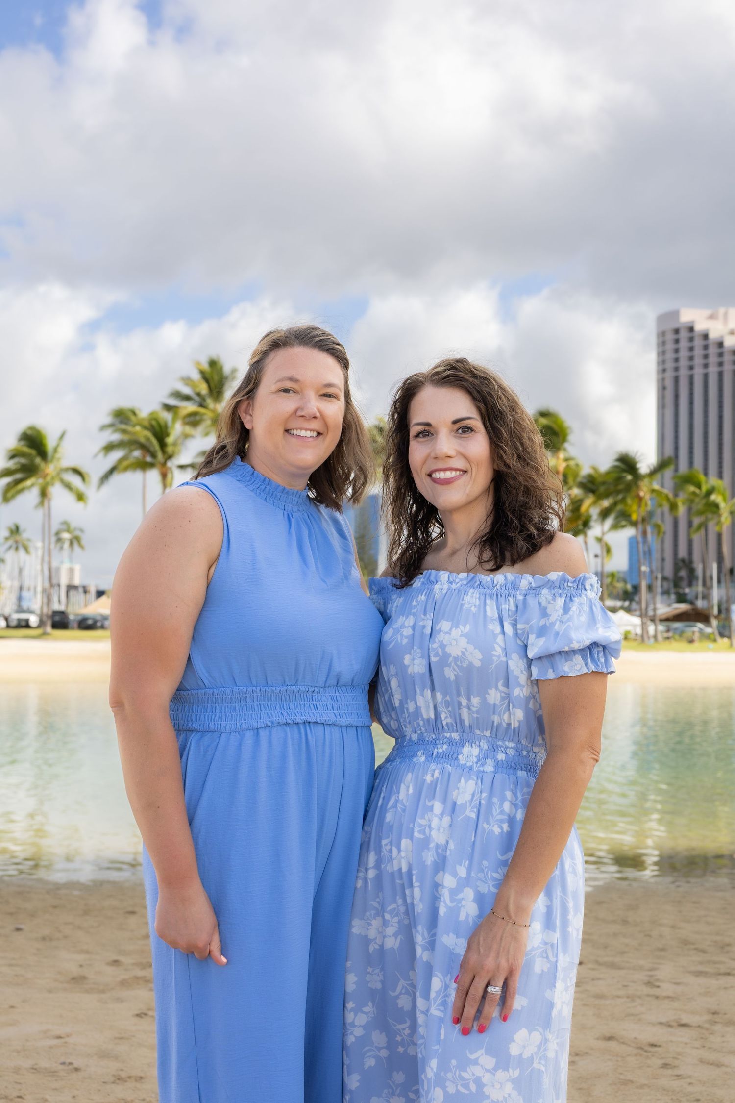 Two women in blue dresses smiling on a Hawaiian beach with palm trees and a hotel in the background.