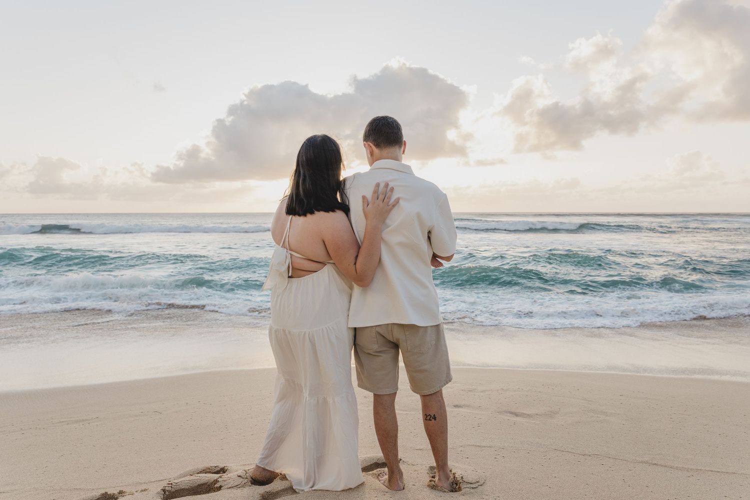 Couple standing together on sandy beach viewed from behind, gazing at ocean waves at sunset.
