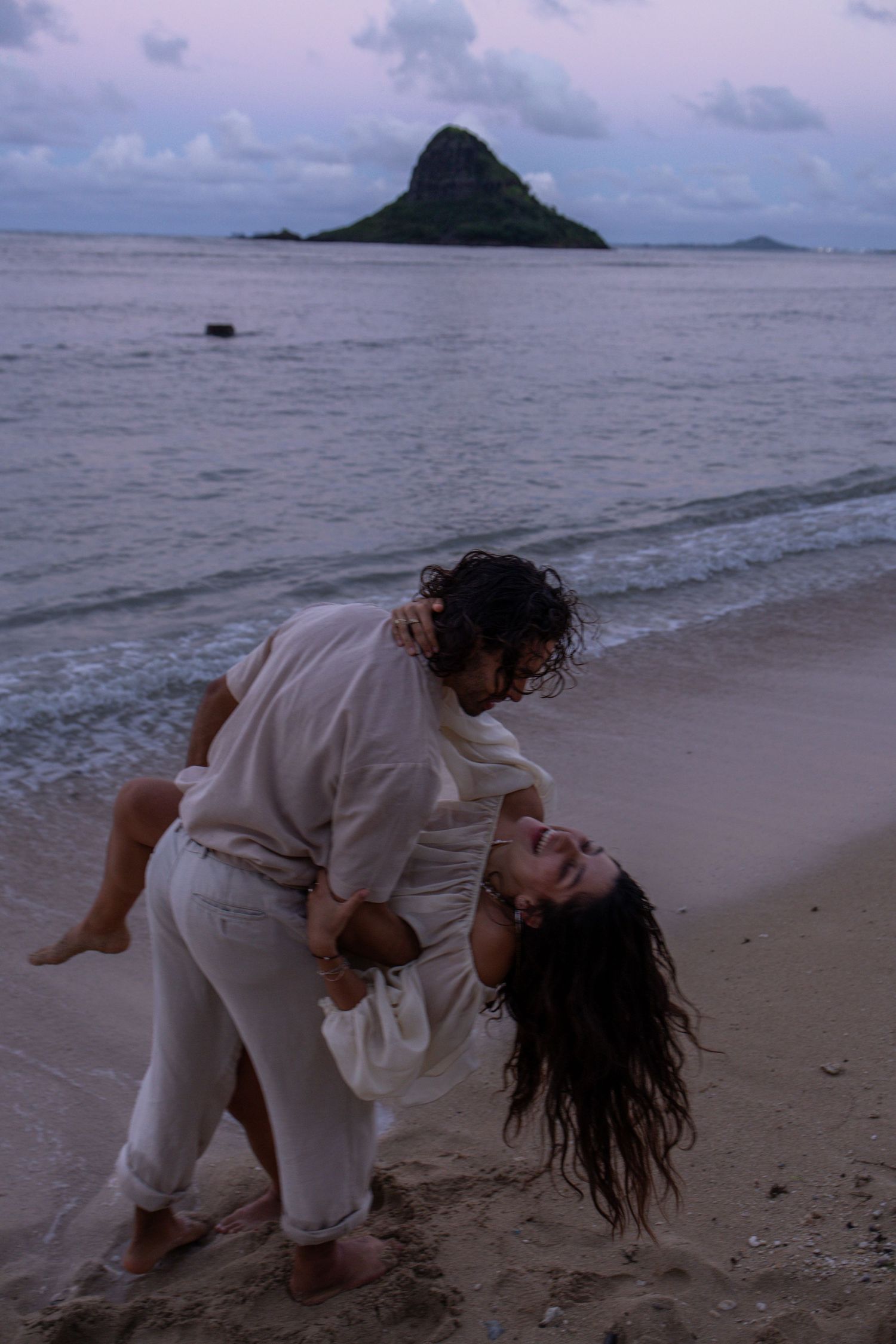 Romantic couple dancing playfully on sandy beach at dusk with Chinaman Hat island backdrop.