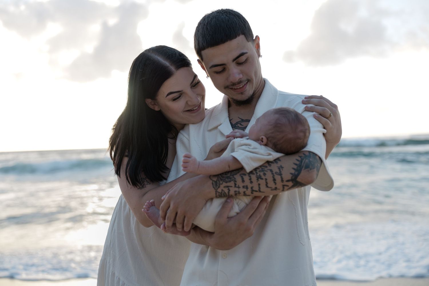 Young couple holding newborn baby together on a beach at sunset in white outfits.