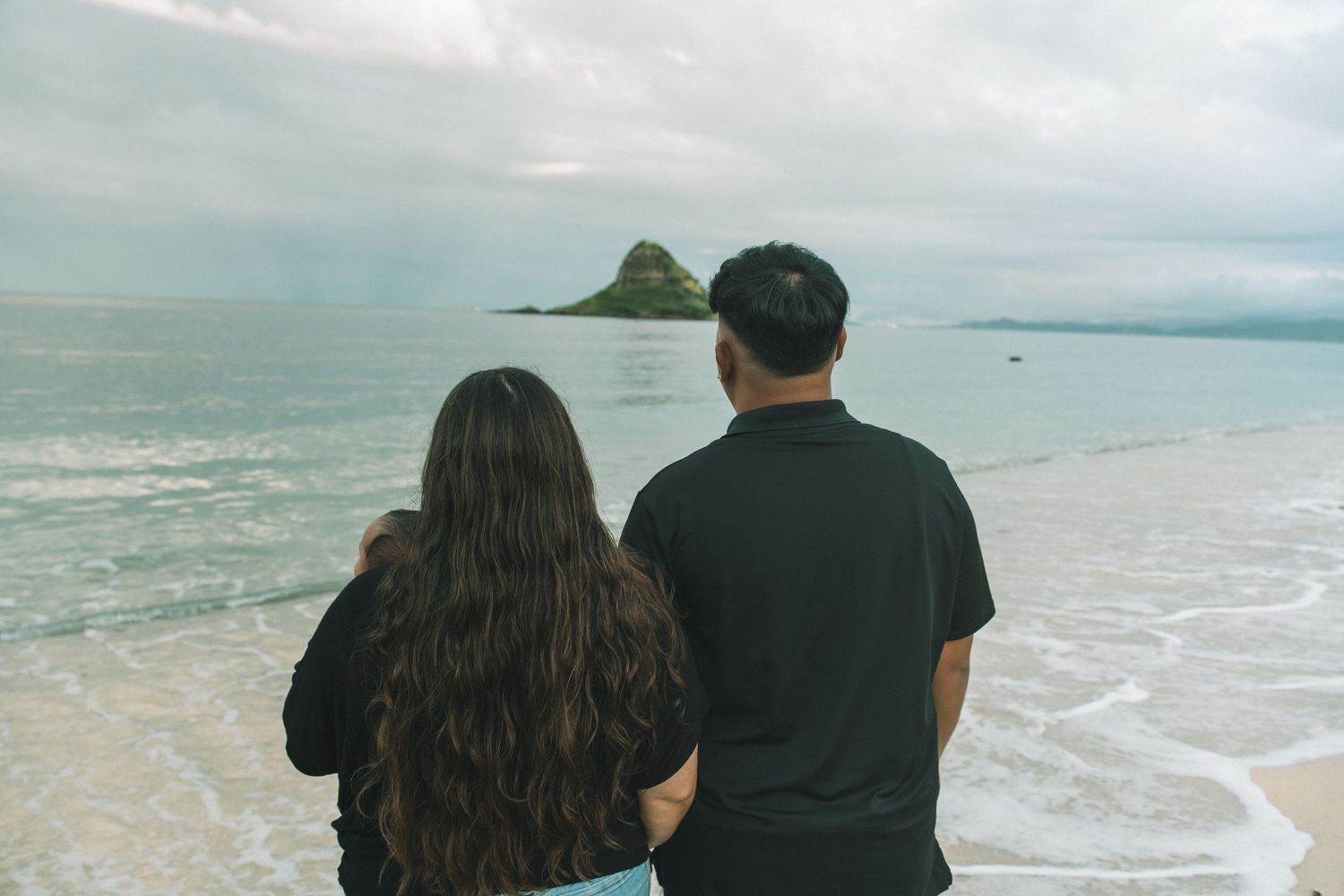A couple stands on a sandy beach gazing at a small island rising from the calm ocean under a cloudy sky.