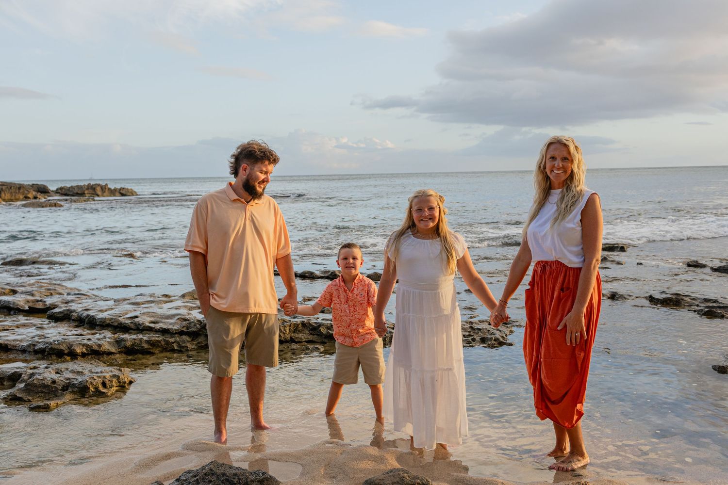 Family of four smiling together on a rocky beach at sunset, parents holding hands with two children.