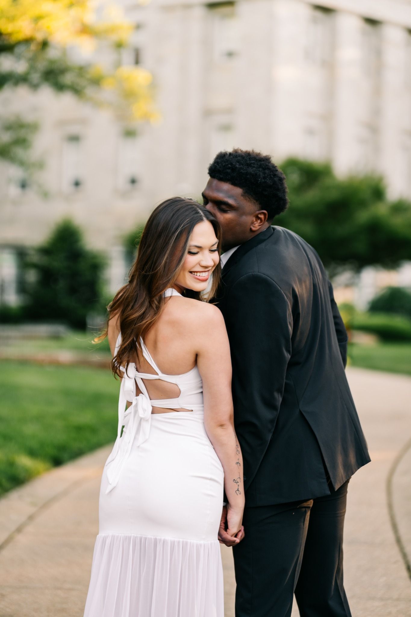 Couple in formal wear holding hands outdoors, woman in white dress smiling as man in black suit leans in close.