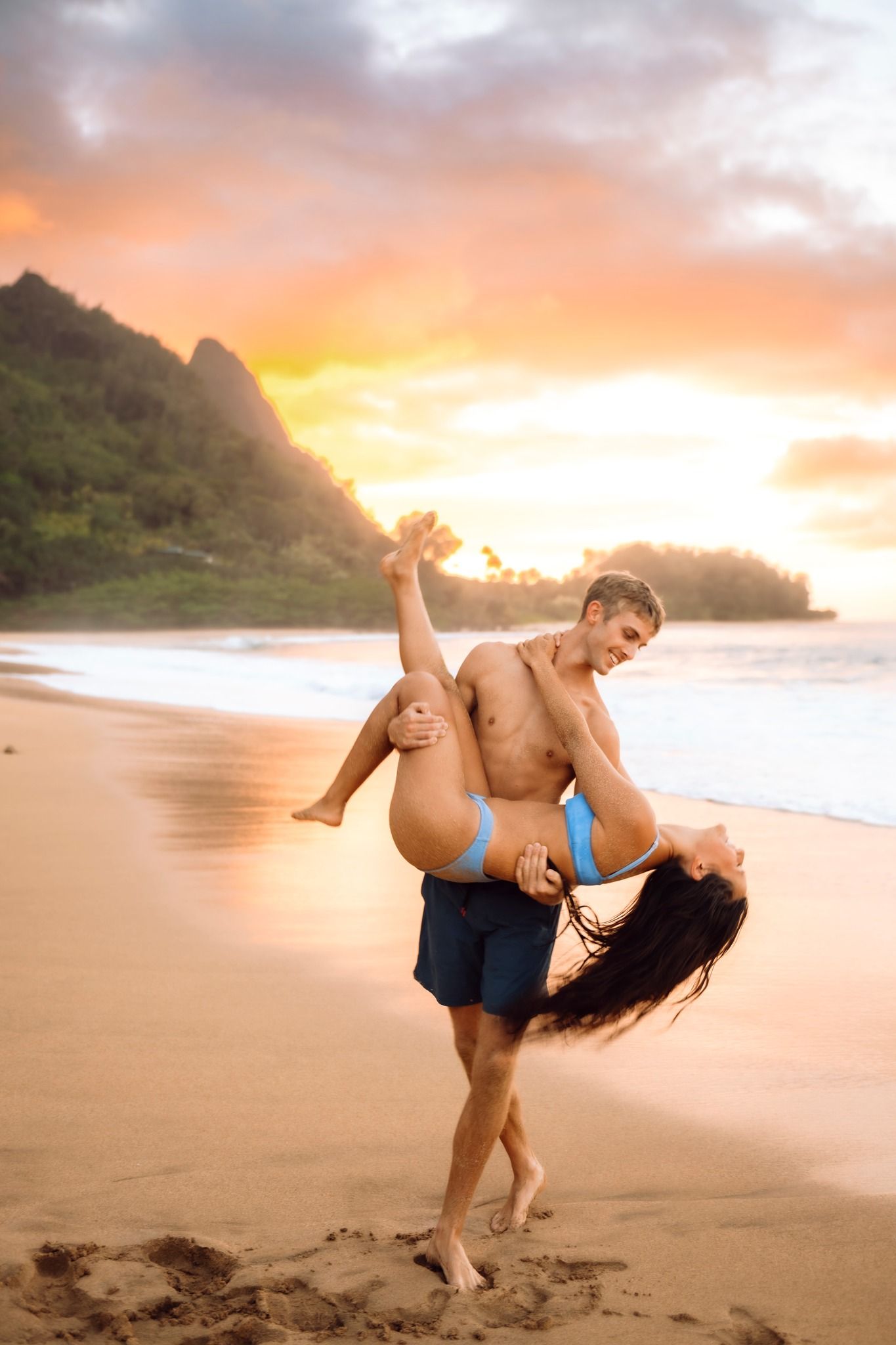 Couple playfully dancing on a tropical beach at sunset, man lifting woman in blue bikini with mountains behind.