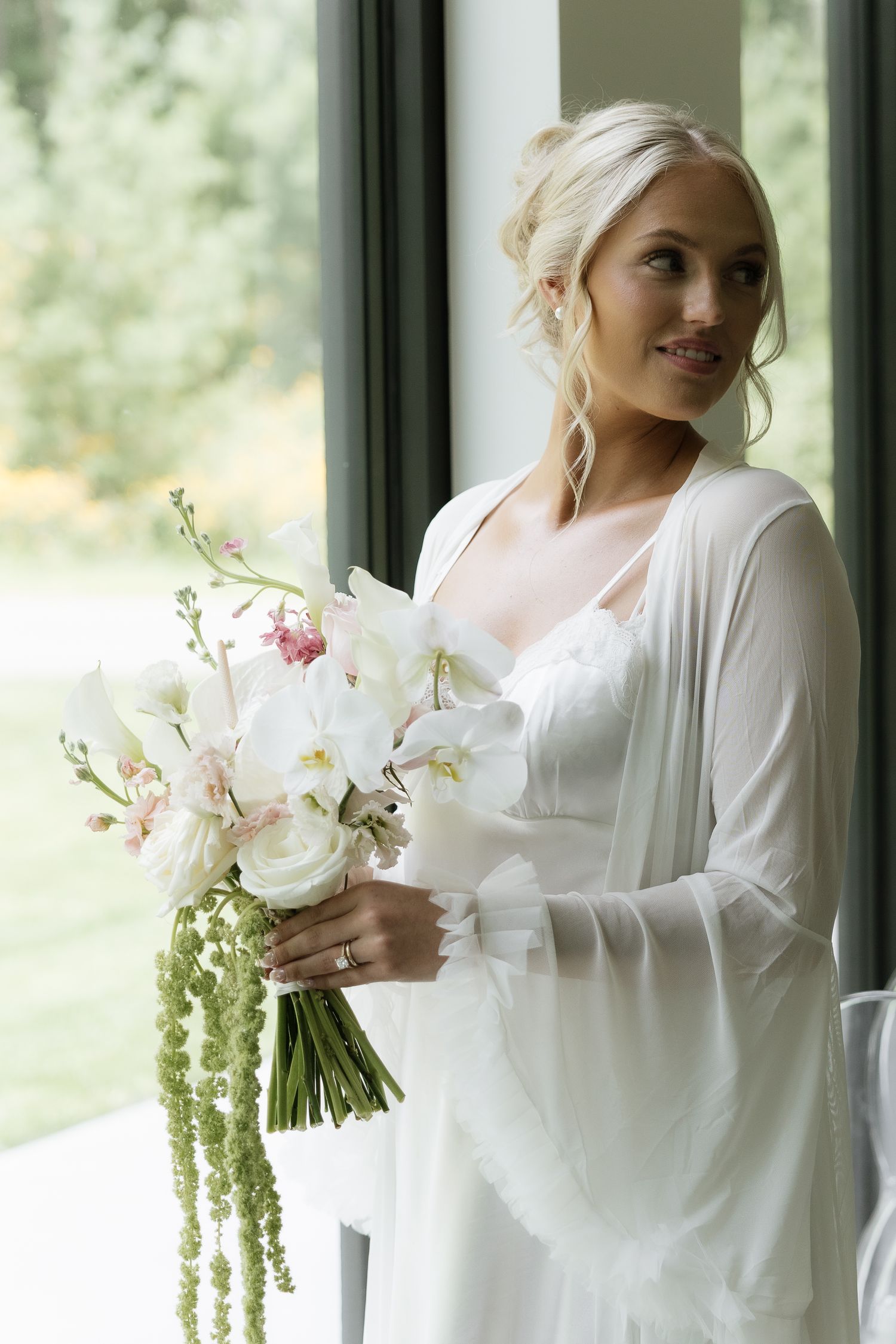 Blonde bride in white robe holding elegant bouquet of white orchids and pink flowers by large window.