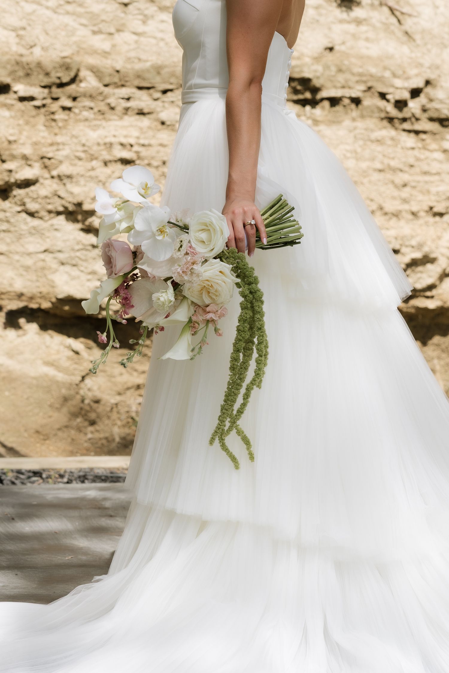 Bride in layered white tulle gown holding cascading bouquet of white and blush flowers with green accents outdoors.
