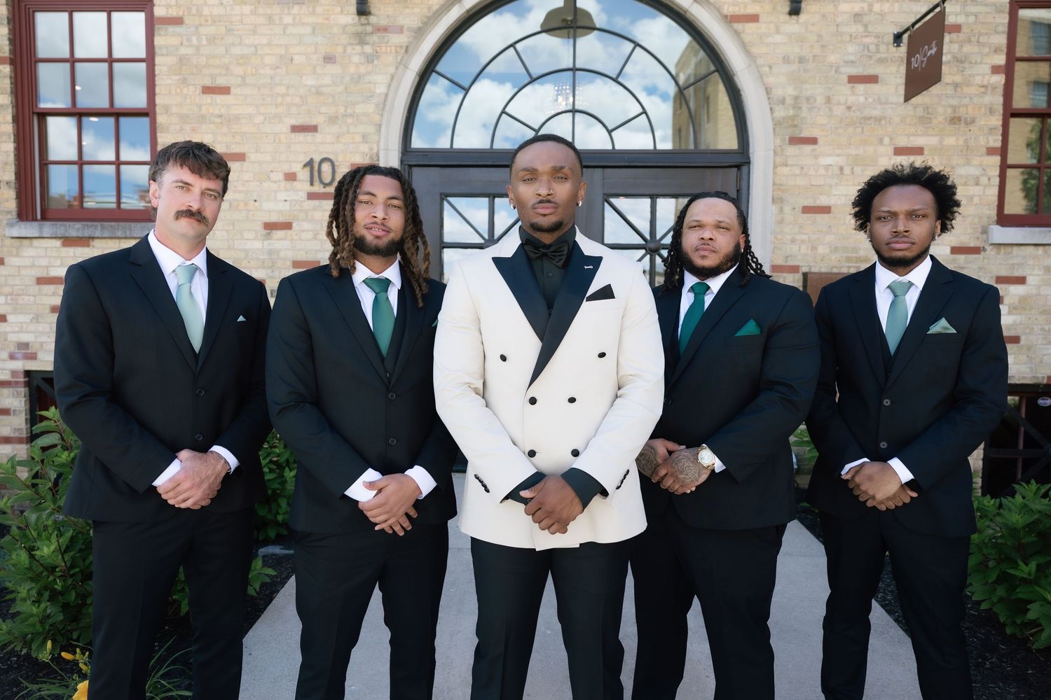 Five men in formal suits with green ties pose outdoors, the center man wearing a white tuxedo jacket.