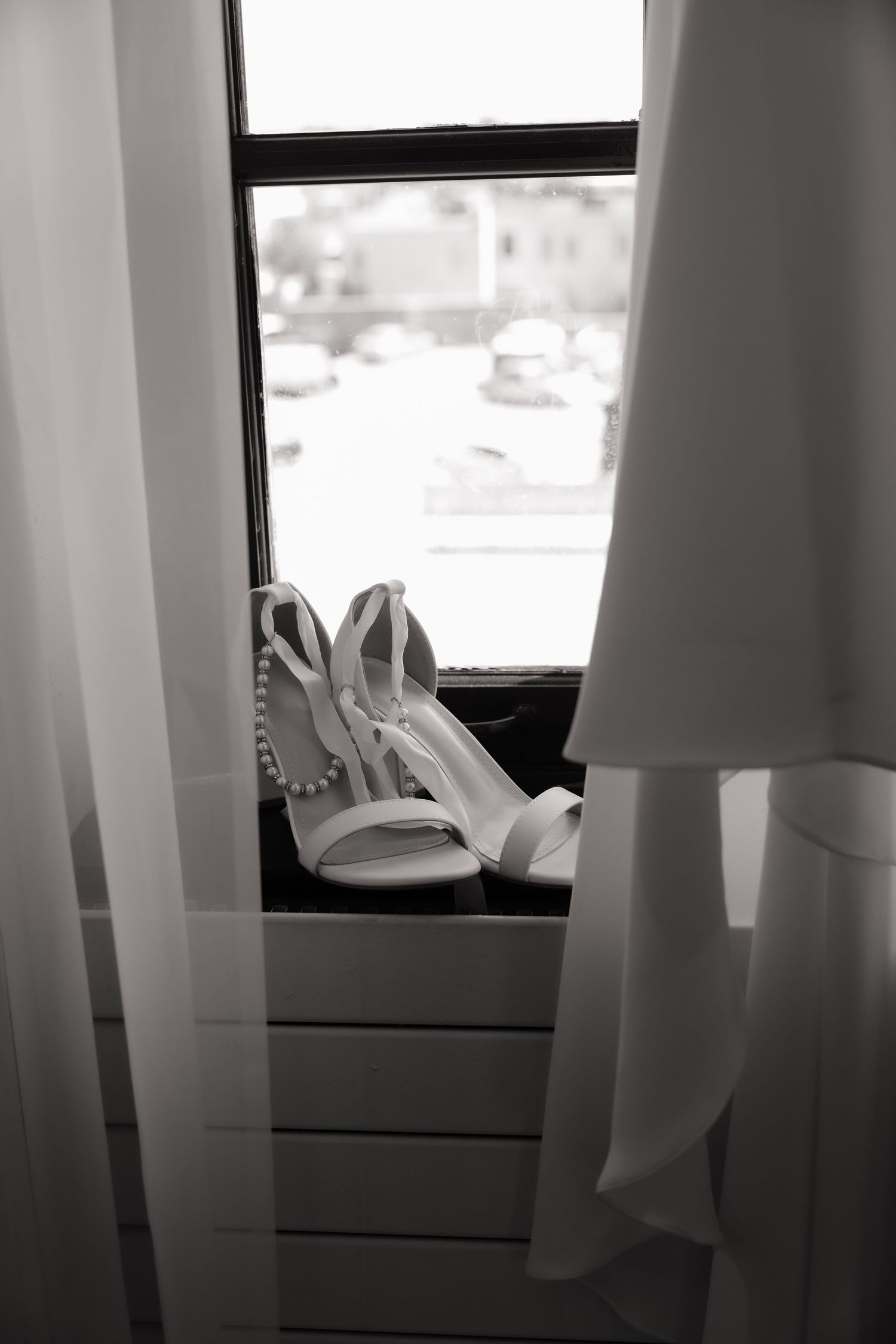 Black and white photo of bridal heels and pearl necklace on windowsill with sheer curtains and wedding dress.