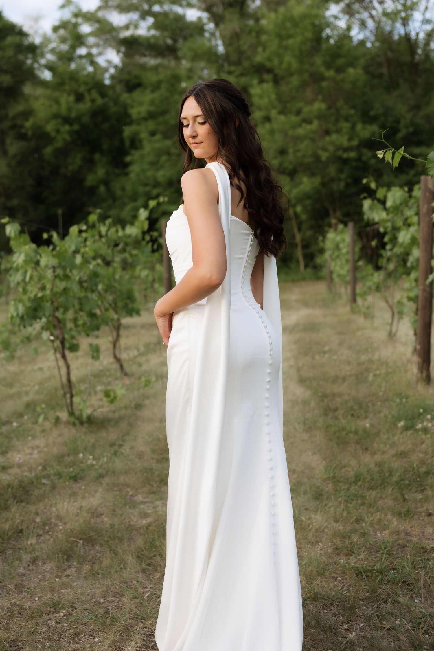 Elegant bride in white button-back gown poses gracefully among vineyard rows at golden hour.
