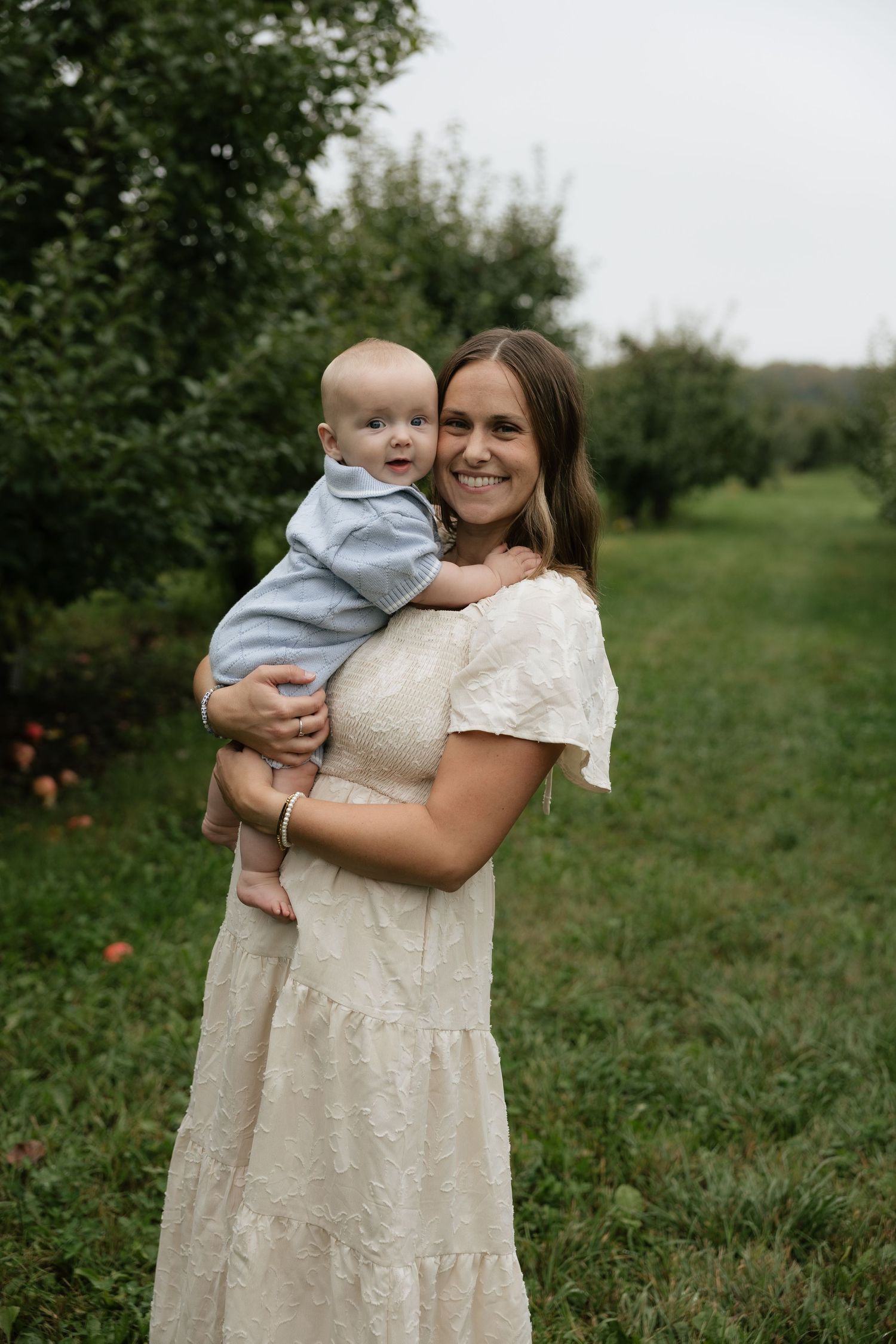 Smiling woman in white dress holding a happy baby in a green orchard setting.