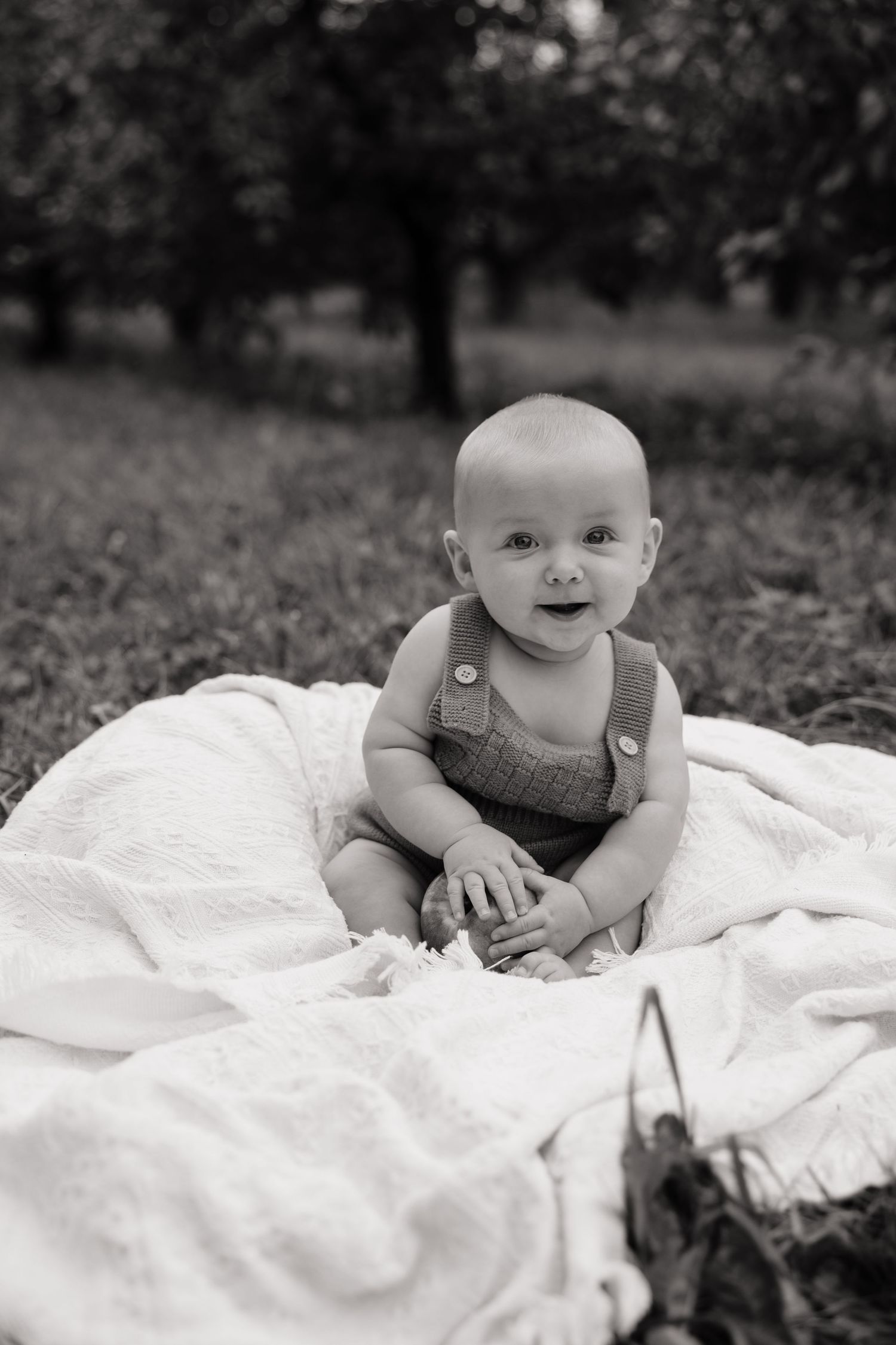 Black and white photo of a happy baby sitting on a white blanket outdoors in a lush green garden.
