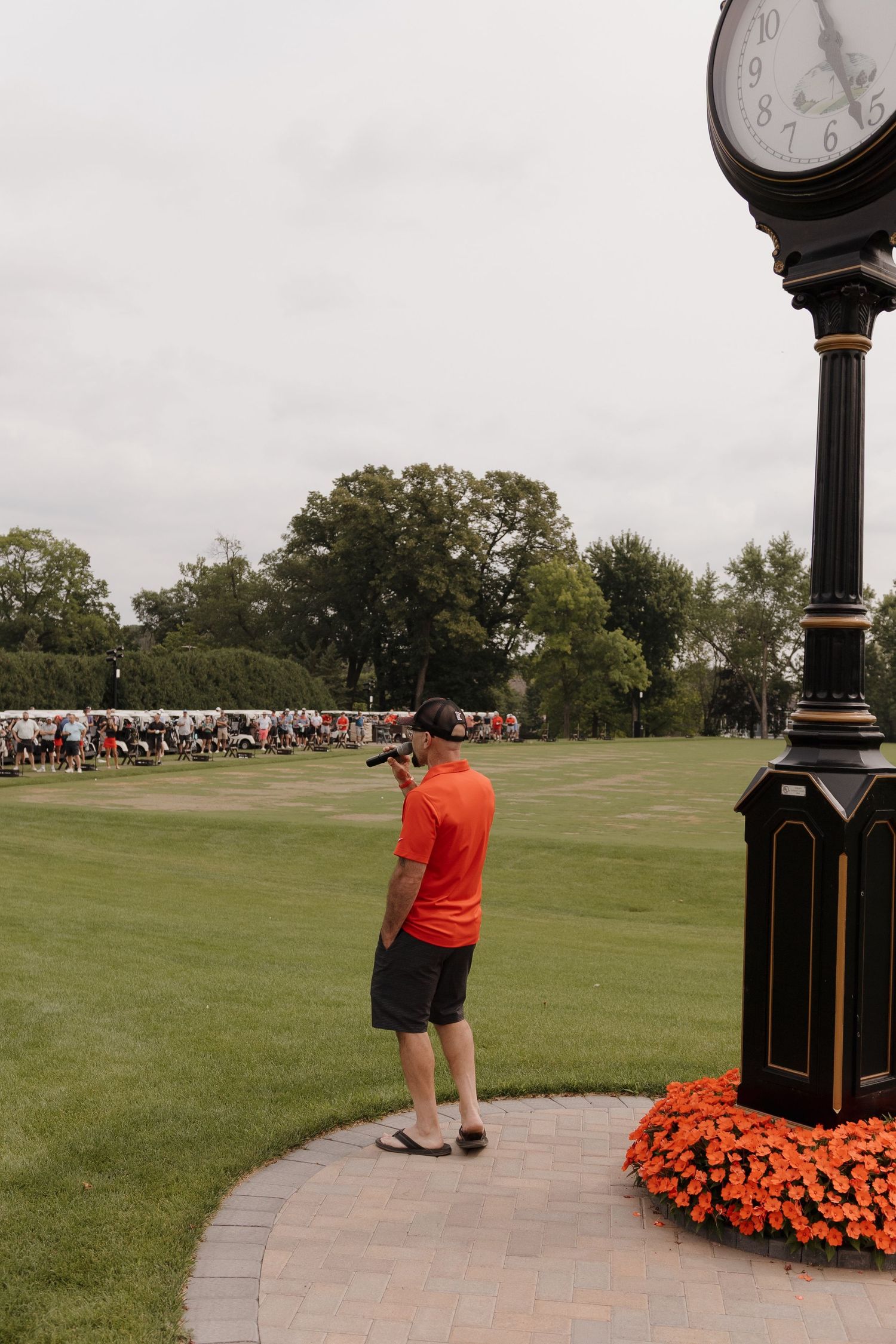 Man in orange shirt photographing a golf course event near a decorative black clock tower with orange flowers.
