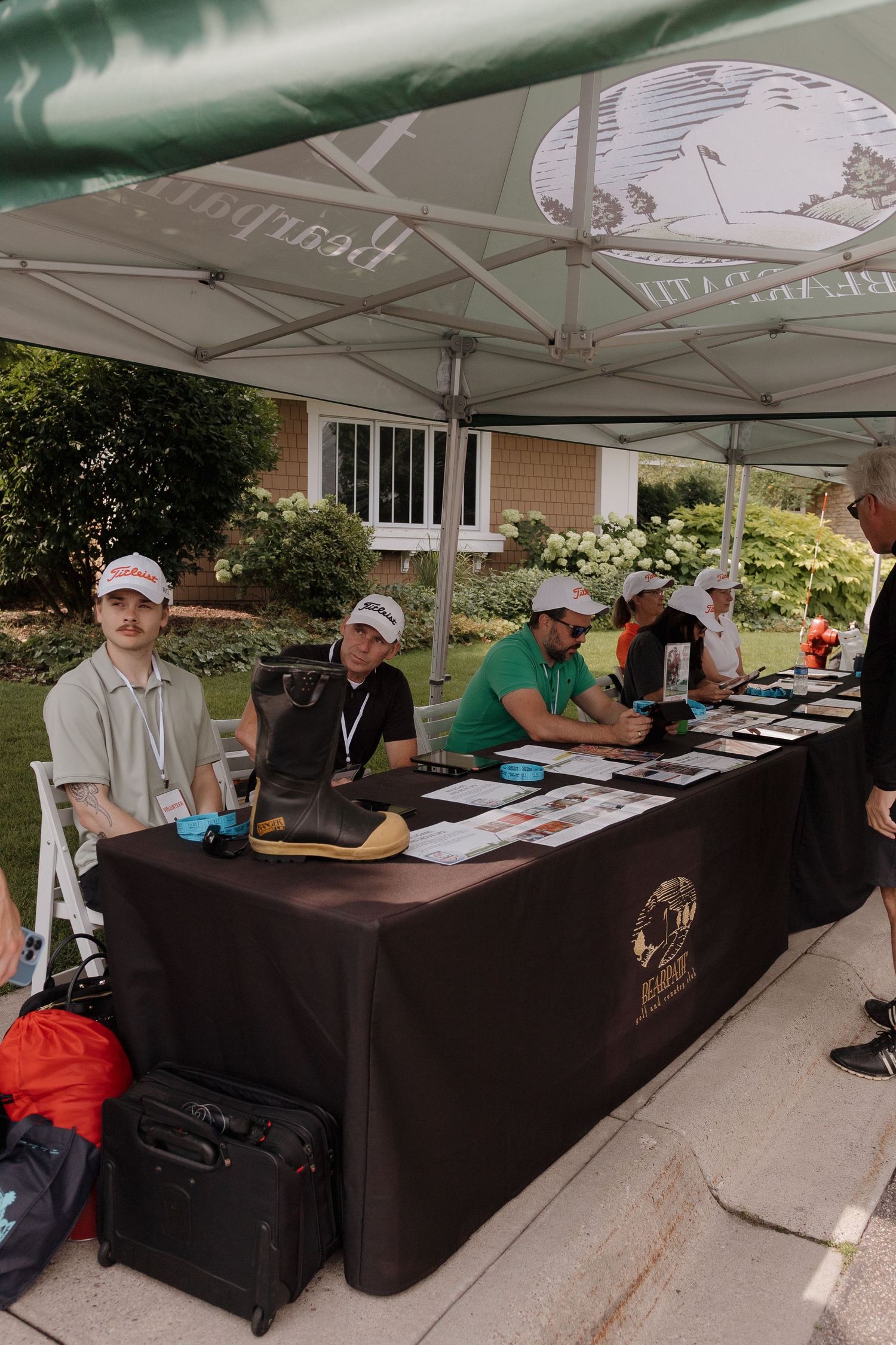 People sitting at a registration table under a white tent outdoors at a golf event with branded tablecloth.