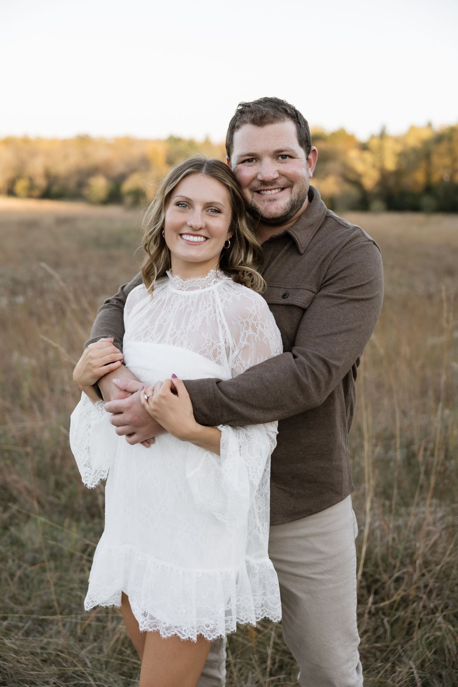 Smiling couple embracing in golden field at sunset, woman in white lace dress, man in brown shirt.