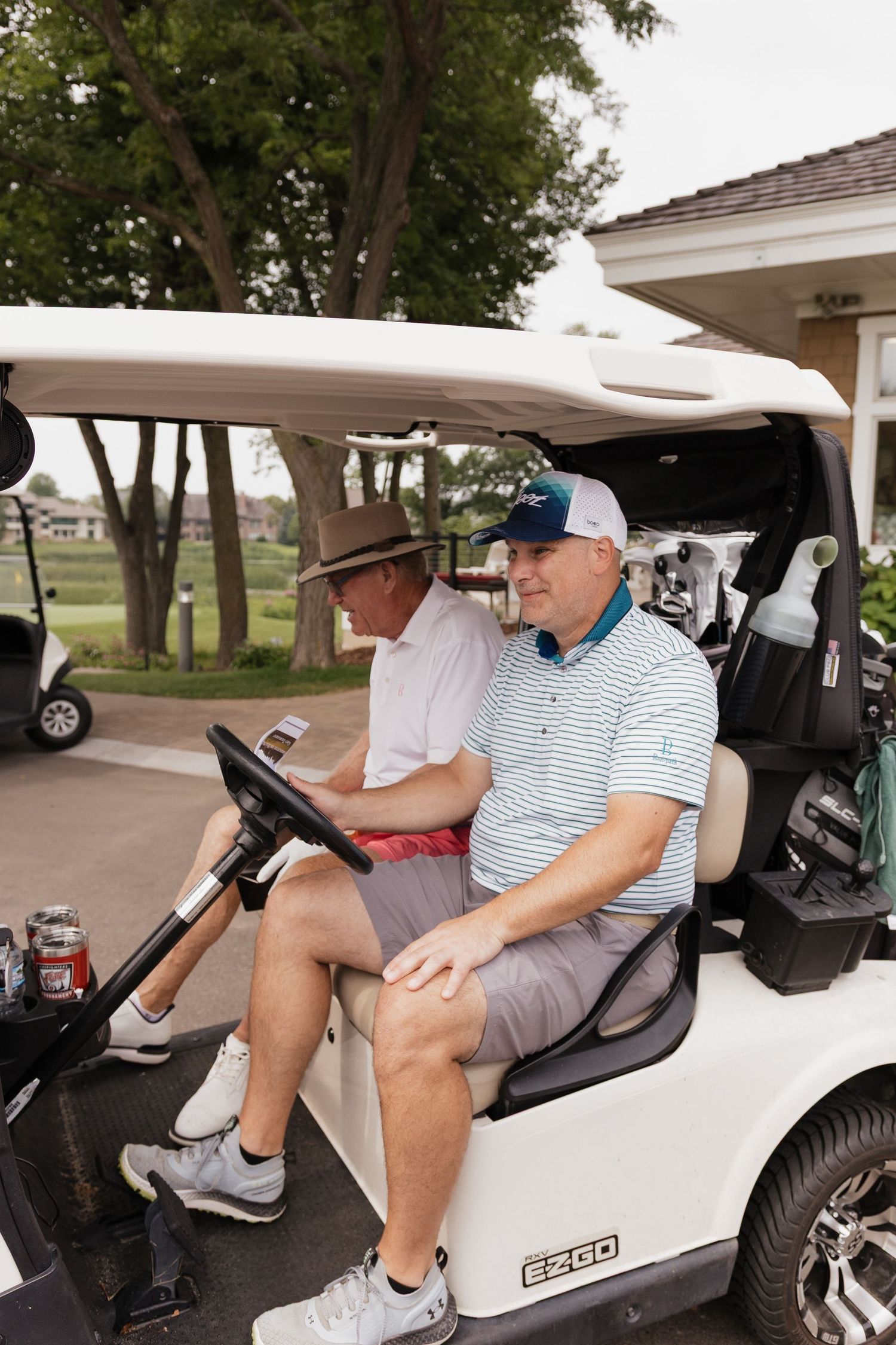 Two senior men in caps sit together in a white E-Z-GO golf cart at a golf course.