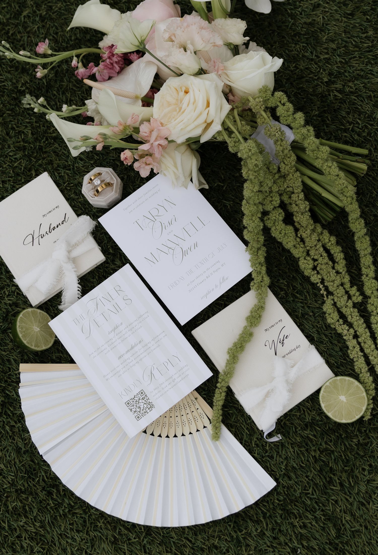 Elegant wedding flat lay with white invitations, floral fan, bouquet of roses, and lime slices on green grass.
