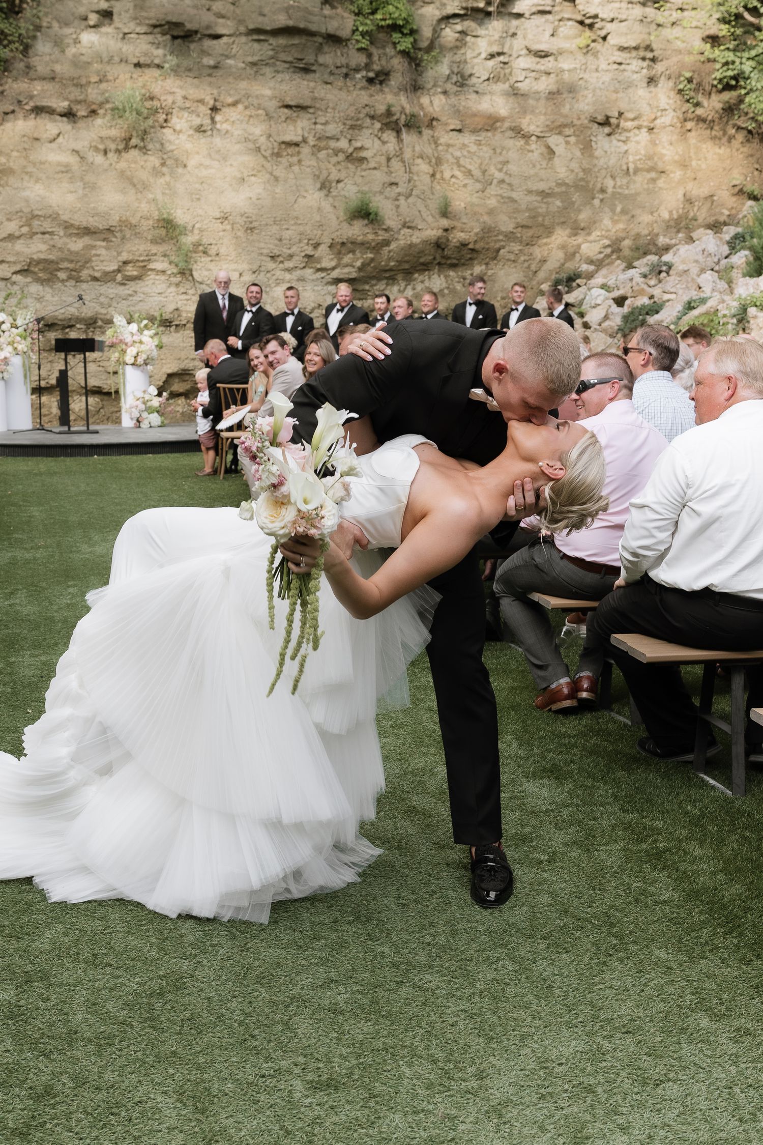 Groom dips bride for romantic kiss after outdoor wedding ceremony while guests watch from wooden benches.
