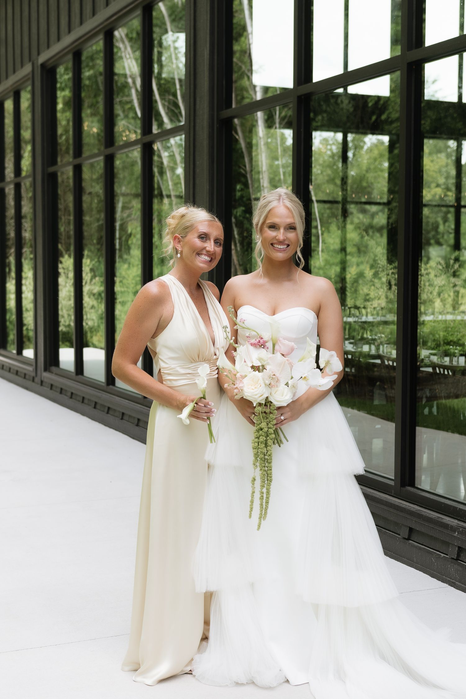 Bride in strapless tiered gown poses with bridesmaid in ivory wrap dress holding floral bouquet.