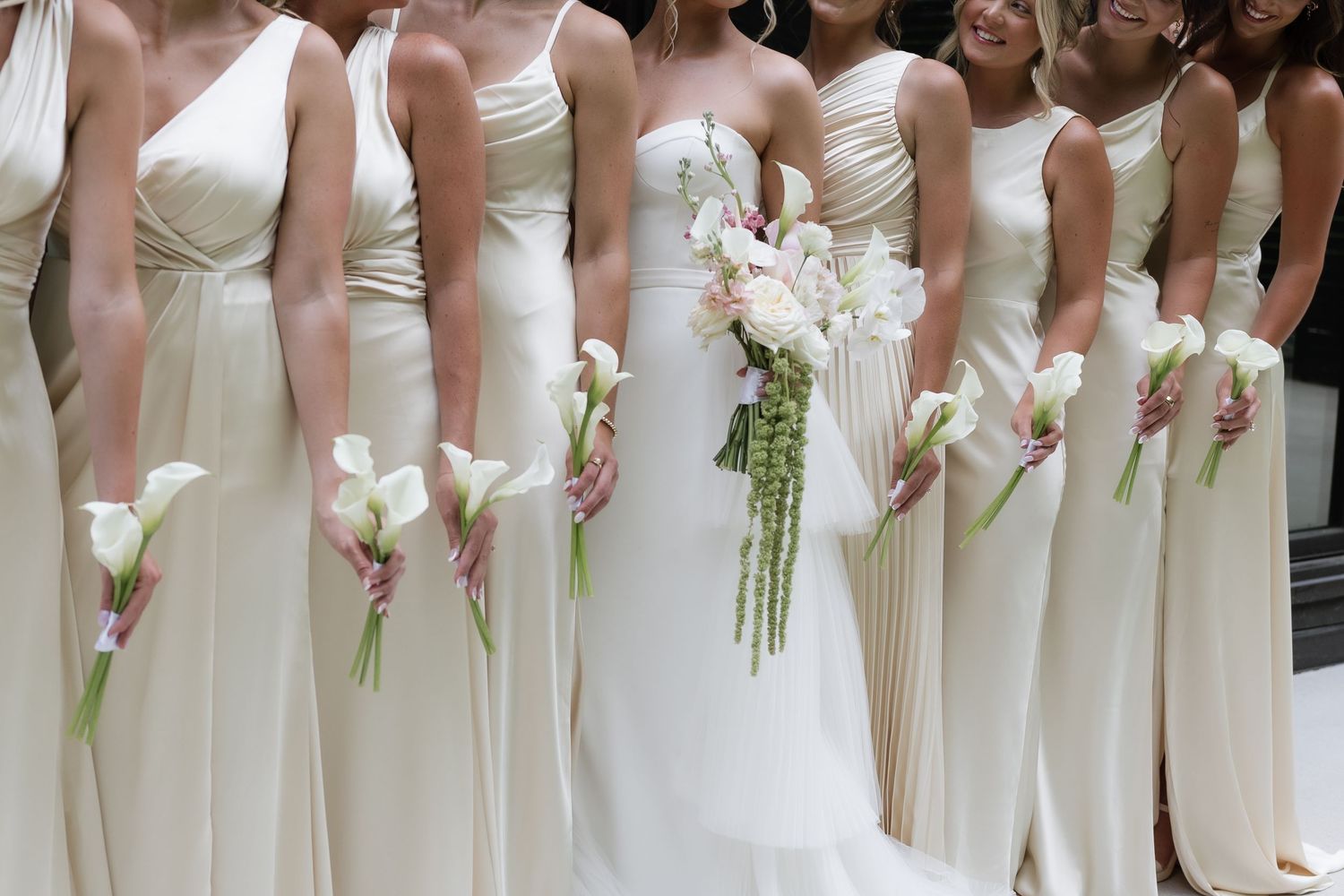 Bride and bridesmaids in white gowns holding calla lily bouquets lined up together at a wedding ceremony.