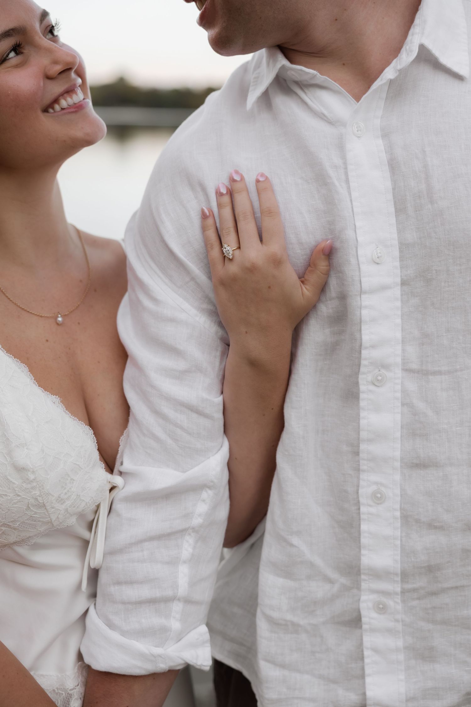 Close-up of womans hand with diamond engagement ring resting on mans white linen shirt by waterfront.