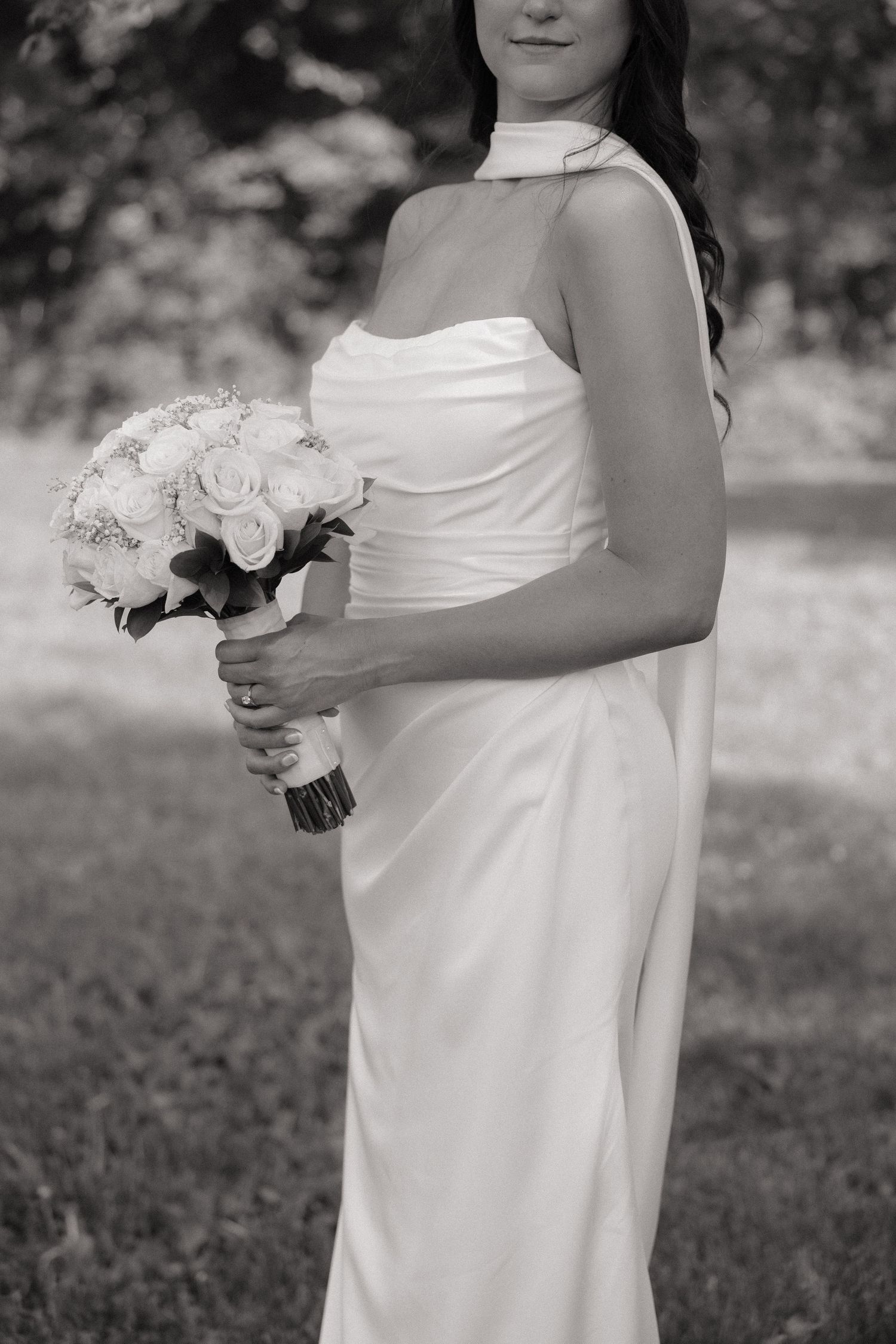 Black and white photo of a bride in a sleek white halter gown holding a bouquet of white roses outdoors.