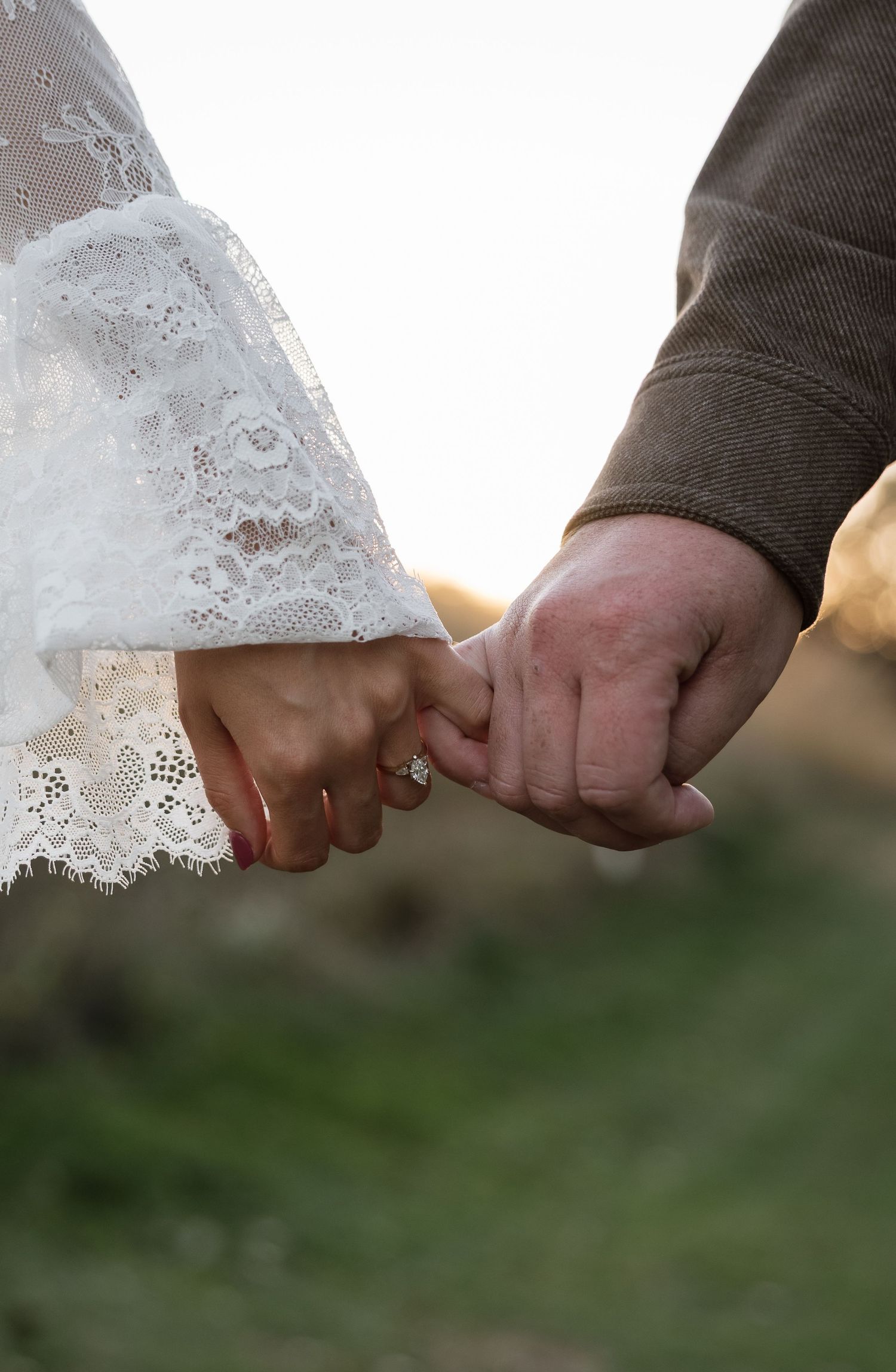 Close-up of a couple holding hands, bride wearing lace wedding dress sleeve with diamond engagement ring outdoors.