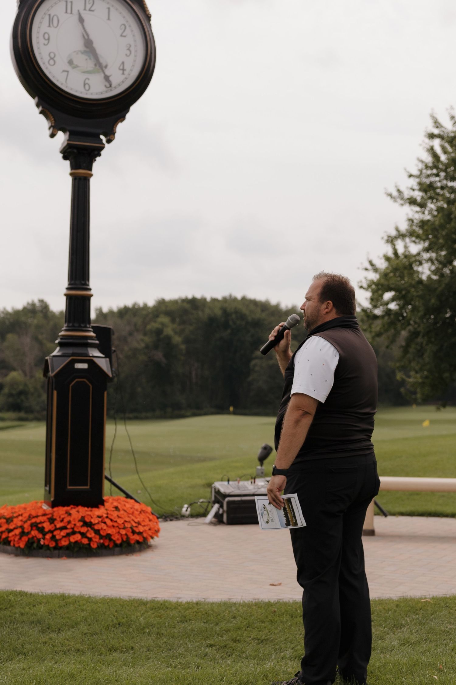 Man speaking into microphone near ornate black clock tower at golf course with orange flowers.