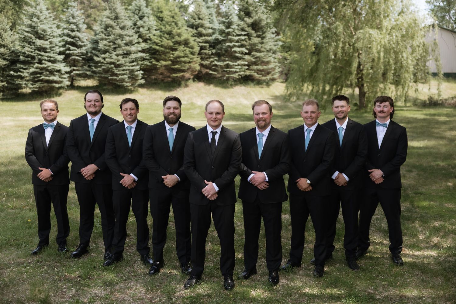 Nine groomsmen in matching black suits with light blue ties pose formally outdoors on wedding day.