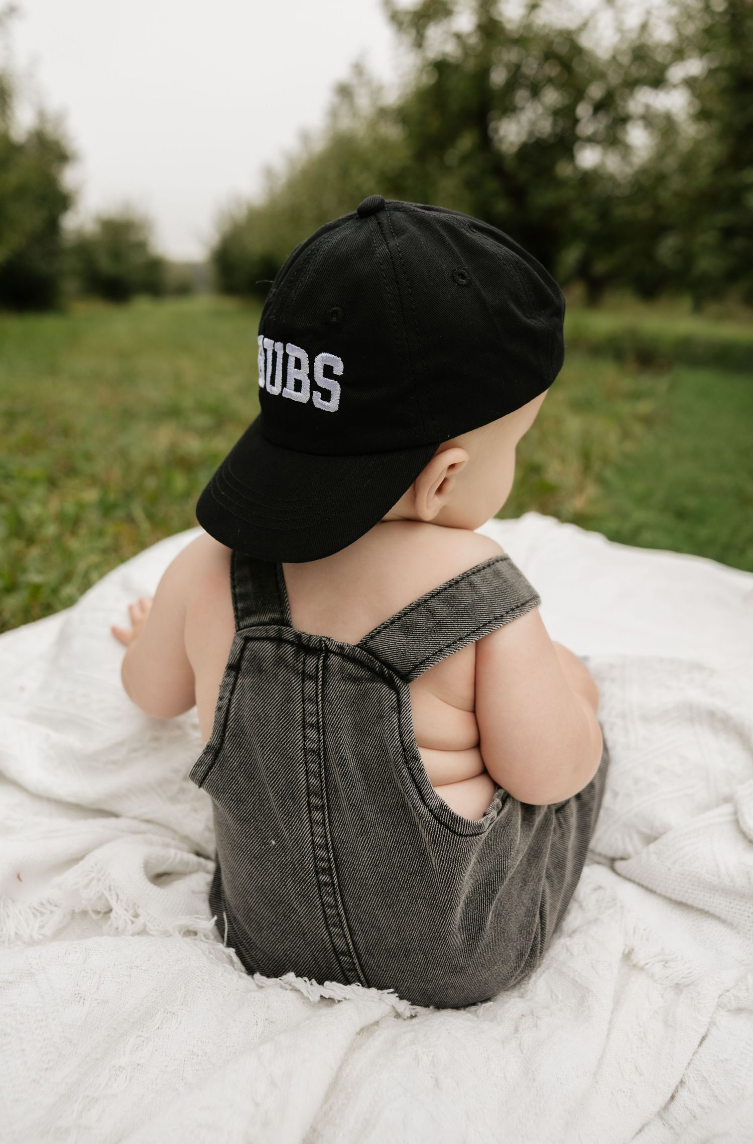 Baby in gray denim overalls and black Cubs cap sits on white blanket in green outdoor setting.