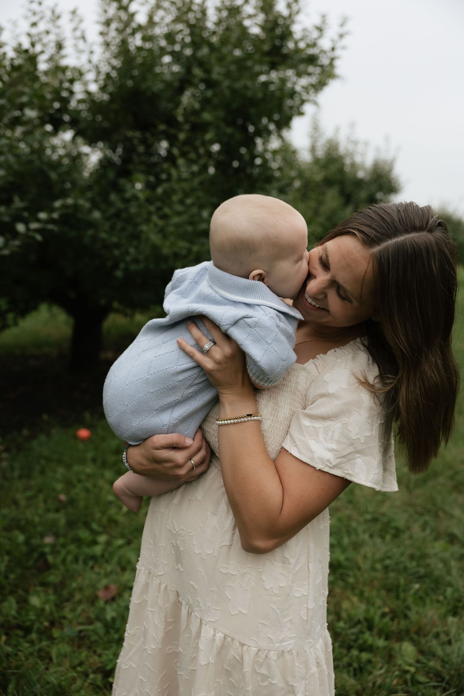 A smiling young mother in a white dress holds her baby in a blue outfit outdoors in a lush green garden.