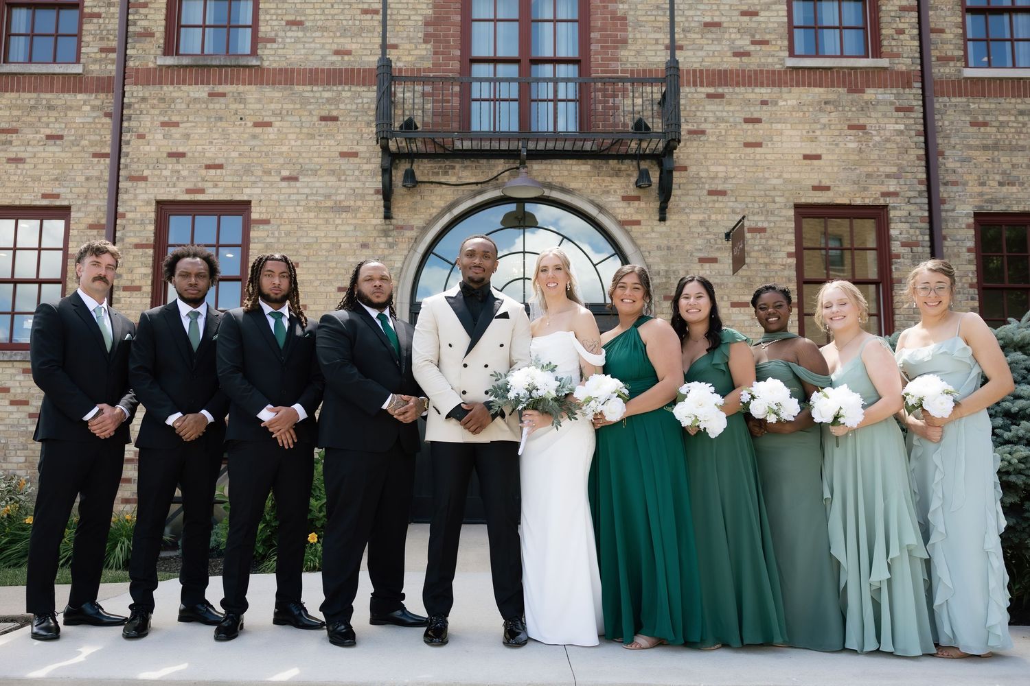 Wedding party poses outside brick venue, bride in white gown, groom in white jacket, groomsmen in black, bridesmaids in green.