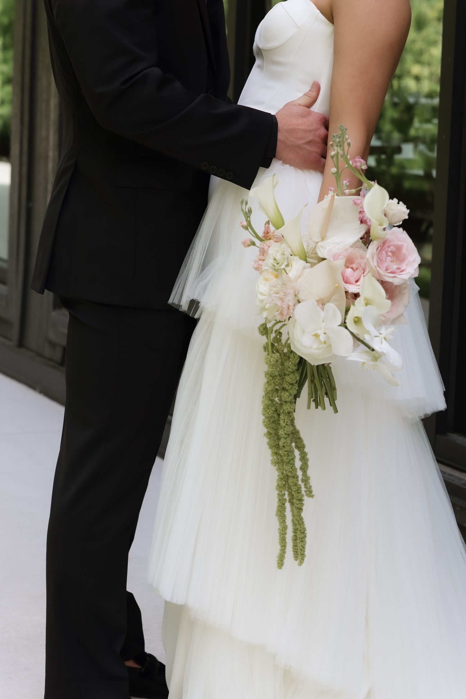 Bride in white tulle gown holding pink and white floral bouquet embraced by groom in black tuxedo.