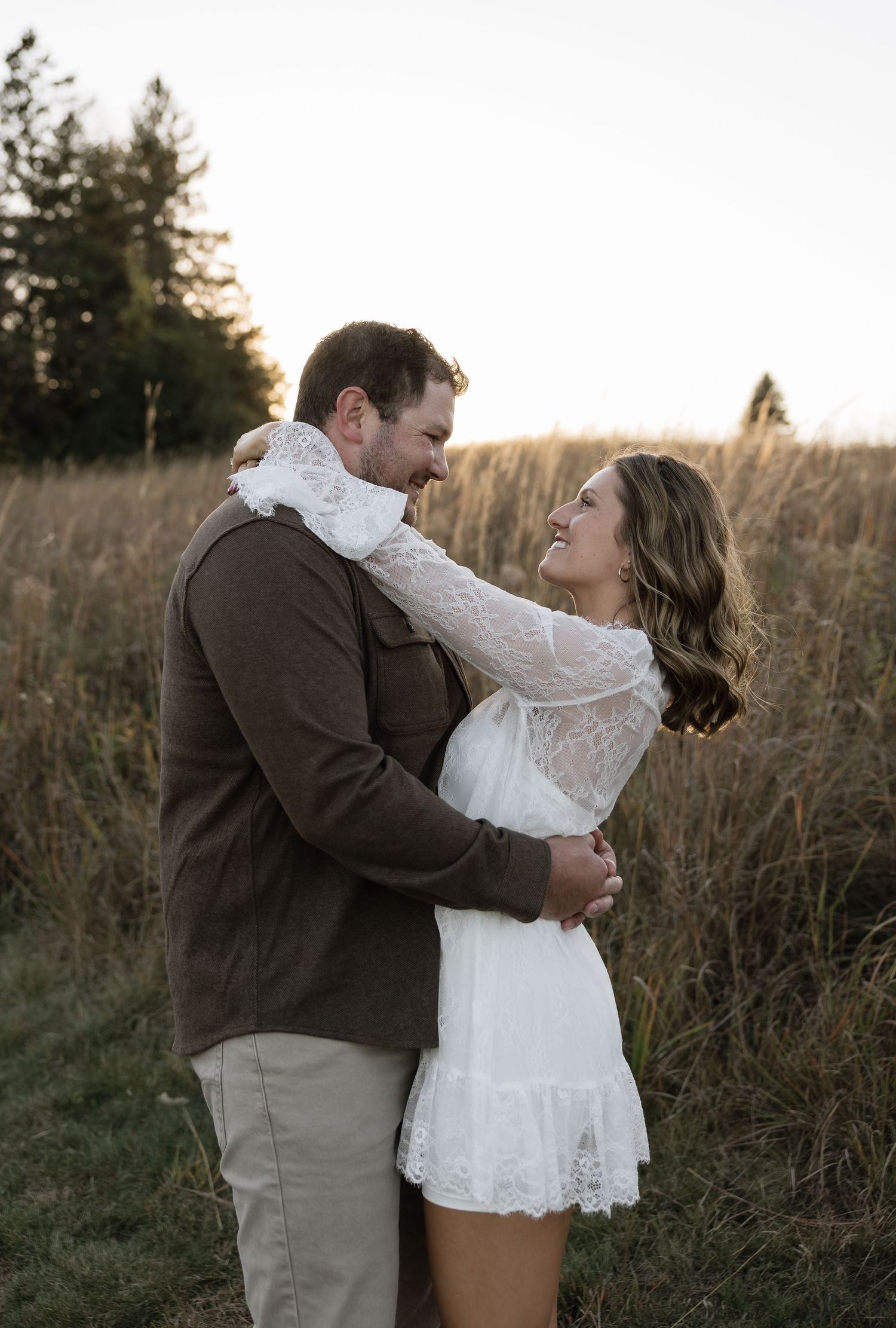 Happy couple embracing in golden field at sunset, woman in white lace dress smiling at man in brown jacket.