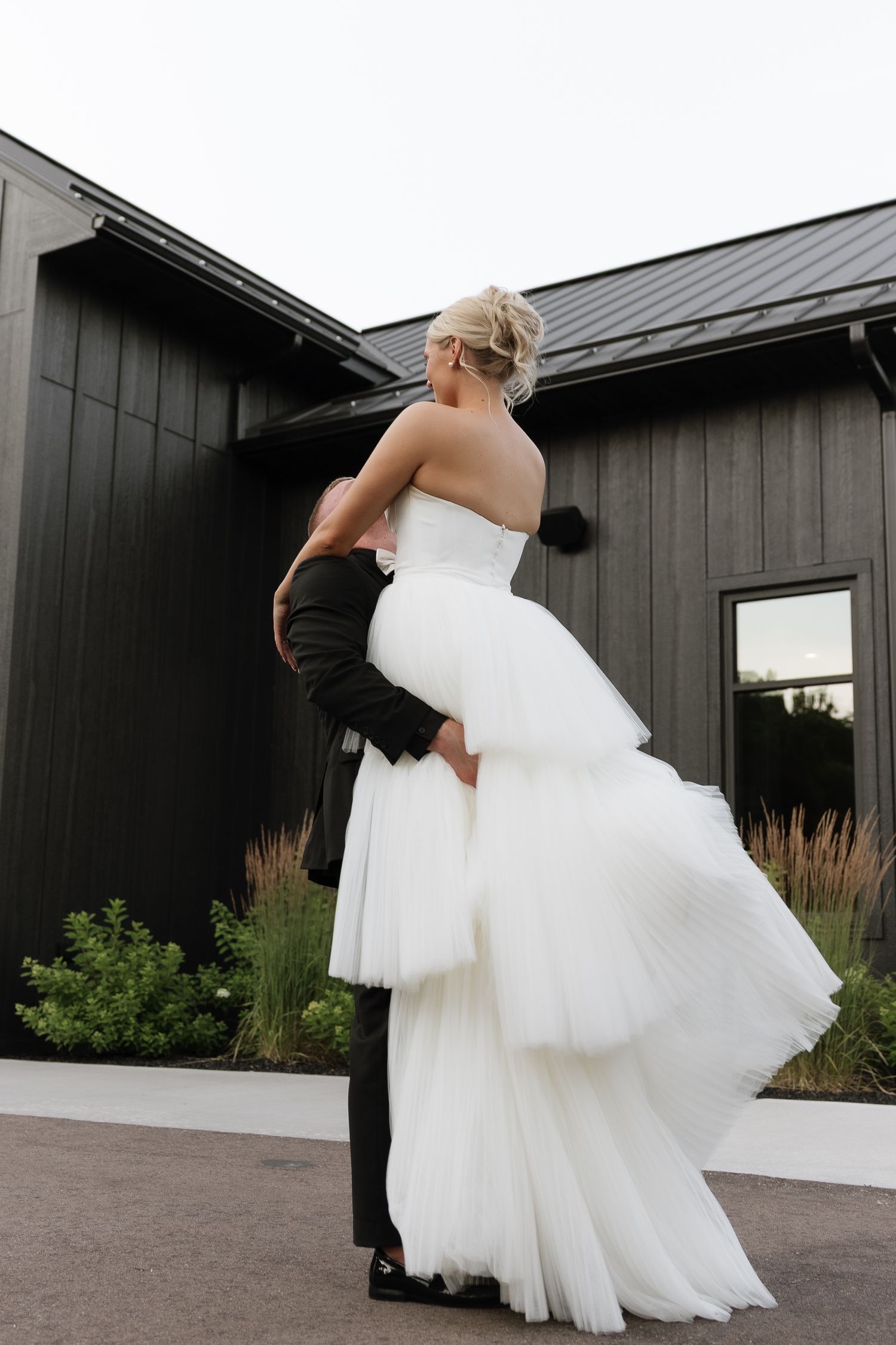 Groom lifts bride in layered tulle wedding gown outside modern black barn wedding venue.