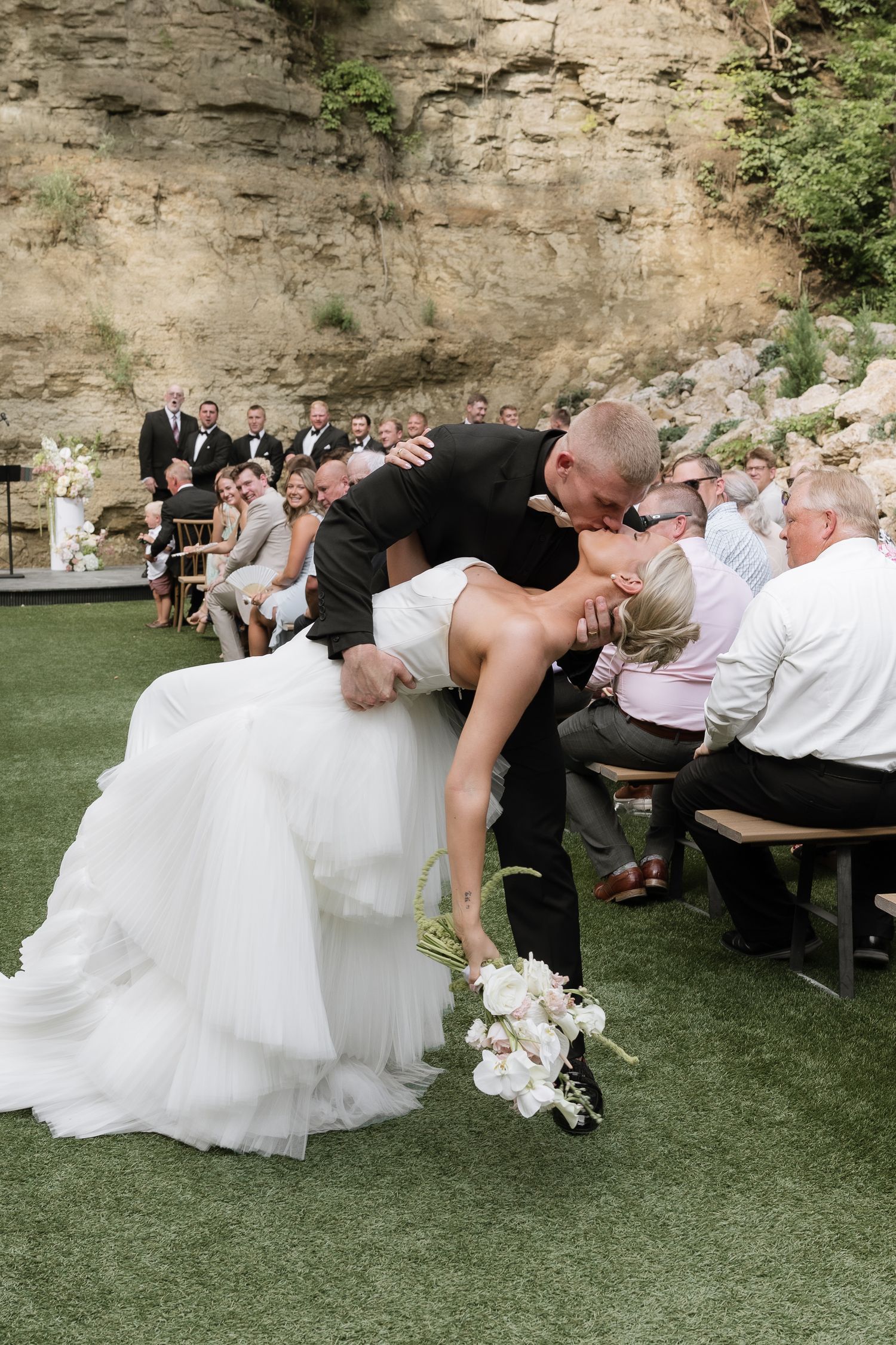 Groom dips bride for a romantic kiss at outdoor wedding ceremony with guests seated beside a rocky cliff backdrop.