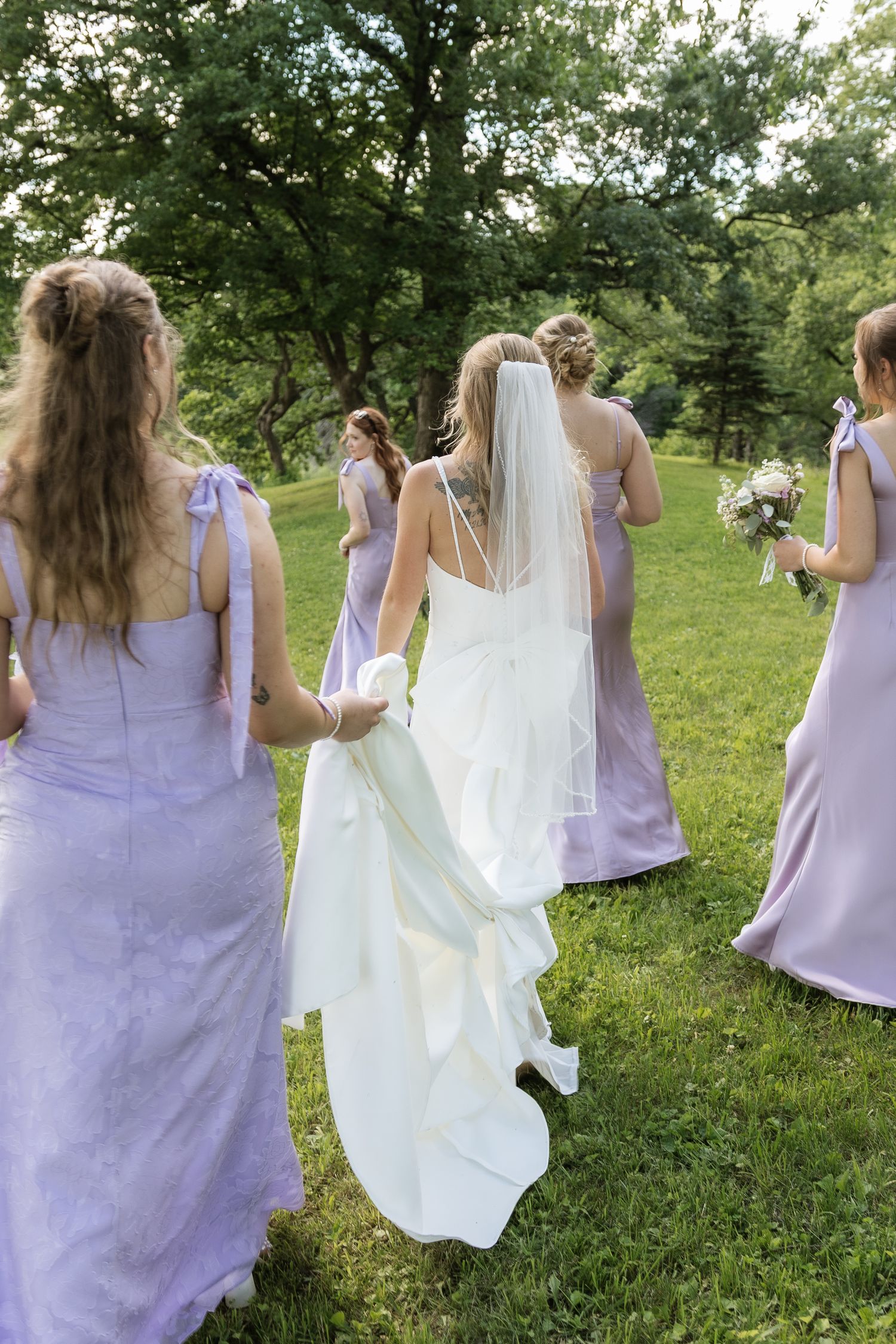 Bride in white gown with veil walks with bridesmaids in lavender dresses across a green lawn.