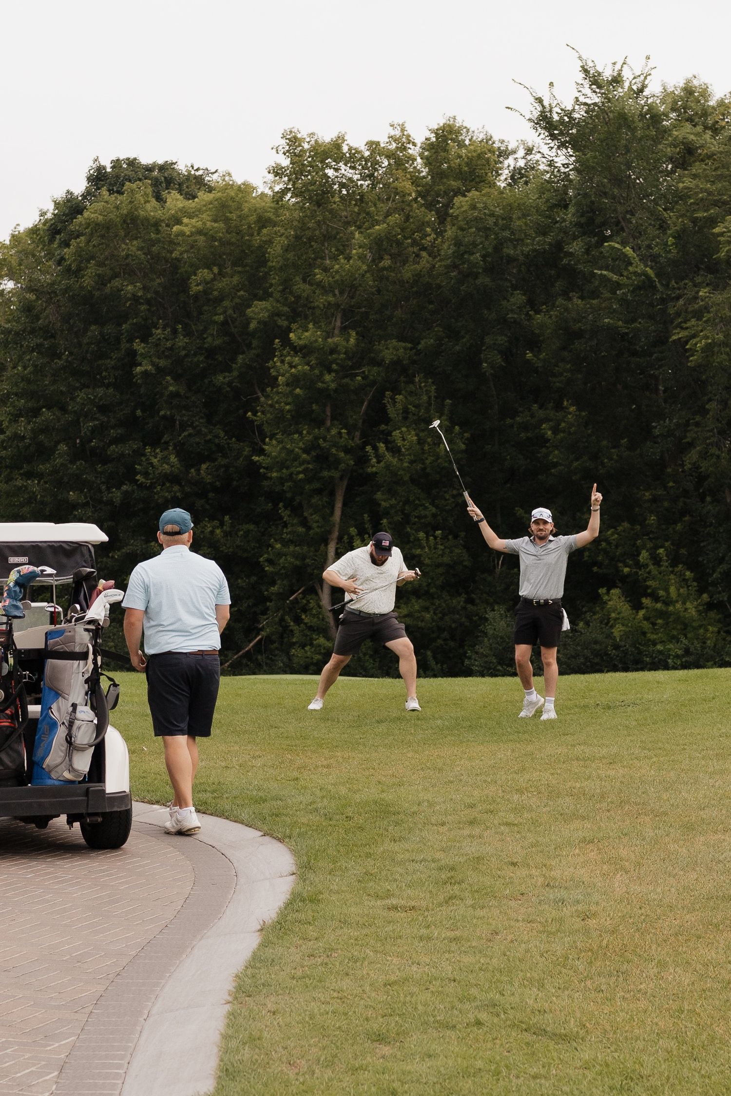 Three men celebrating on a golf course near a golf cart, with one raising his club in triumph on a green fairway.