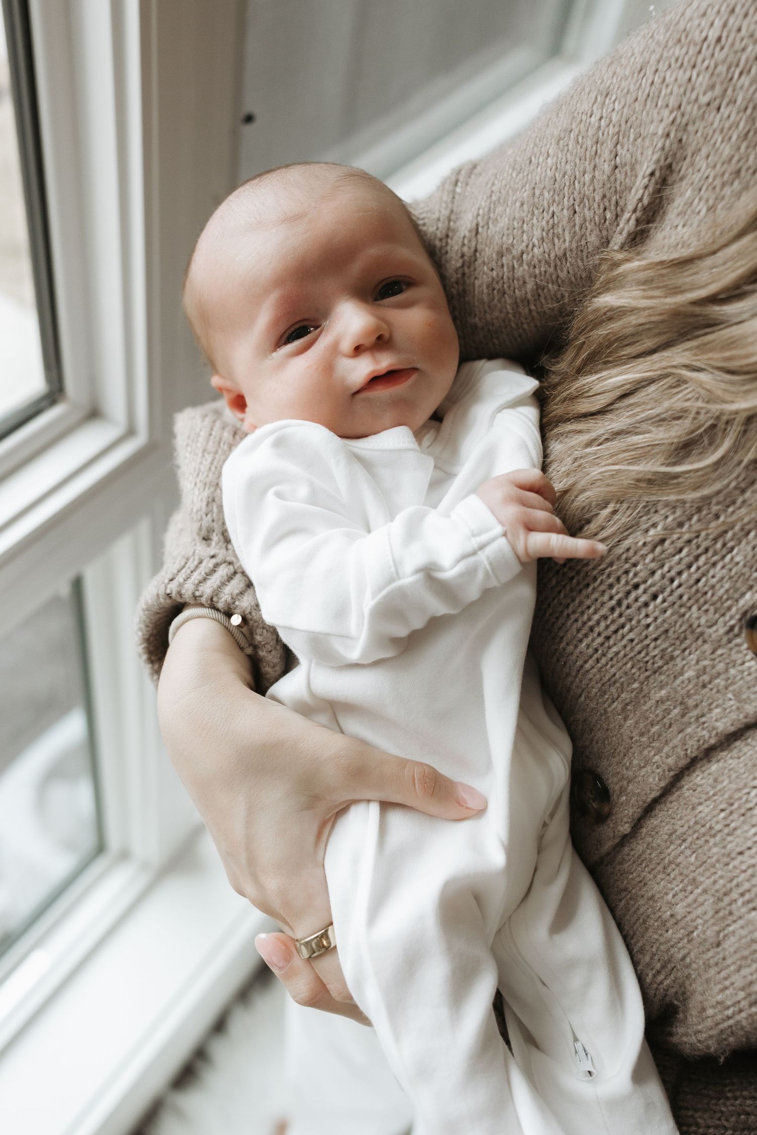 Newborn baby in white sleepsuit being cradled by a parent near a bright window.