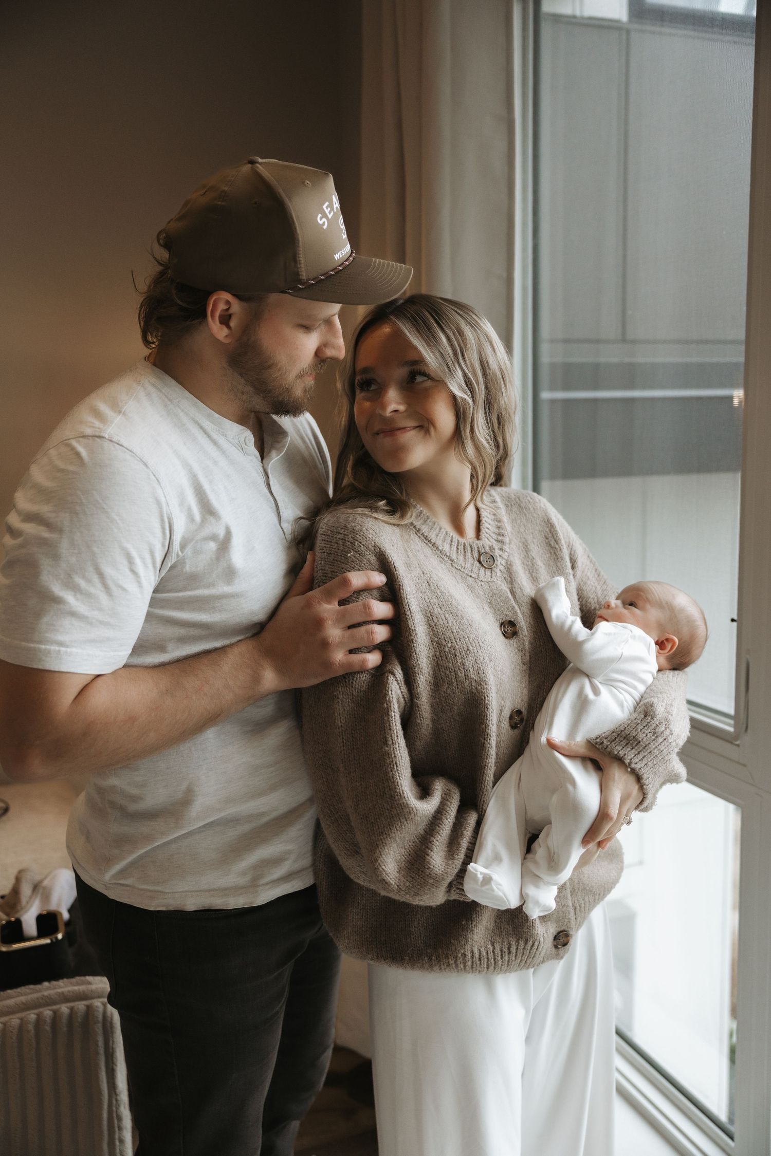 Young couple sharing a tender moment by a window while lovingly holding their newborn baby.
