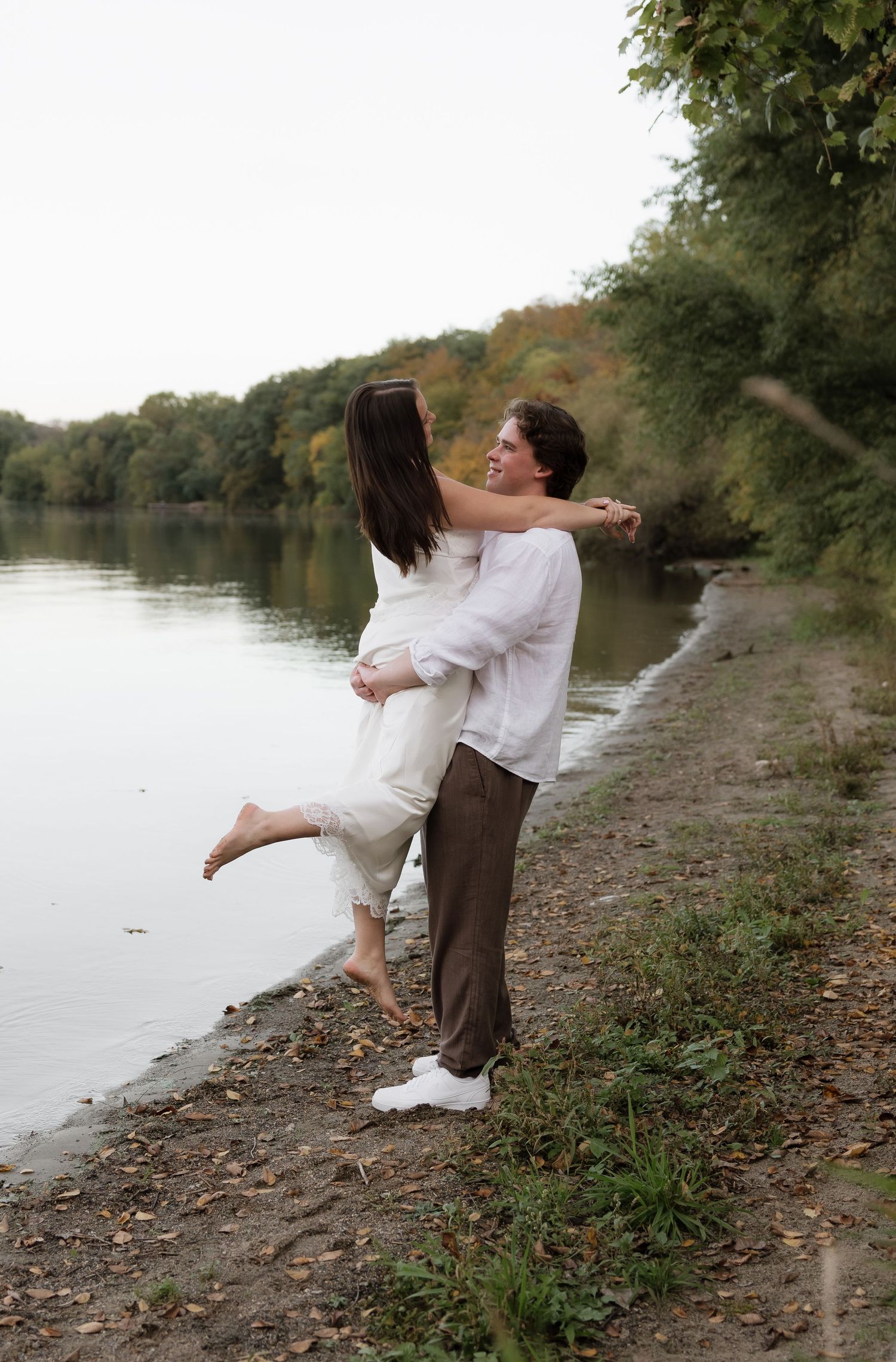 Man joyfully lifting woman in white dress along a scenic lakeside shore during engagement photoshoot.