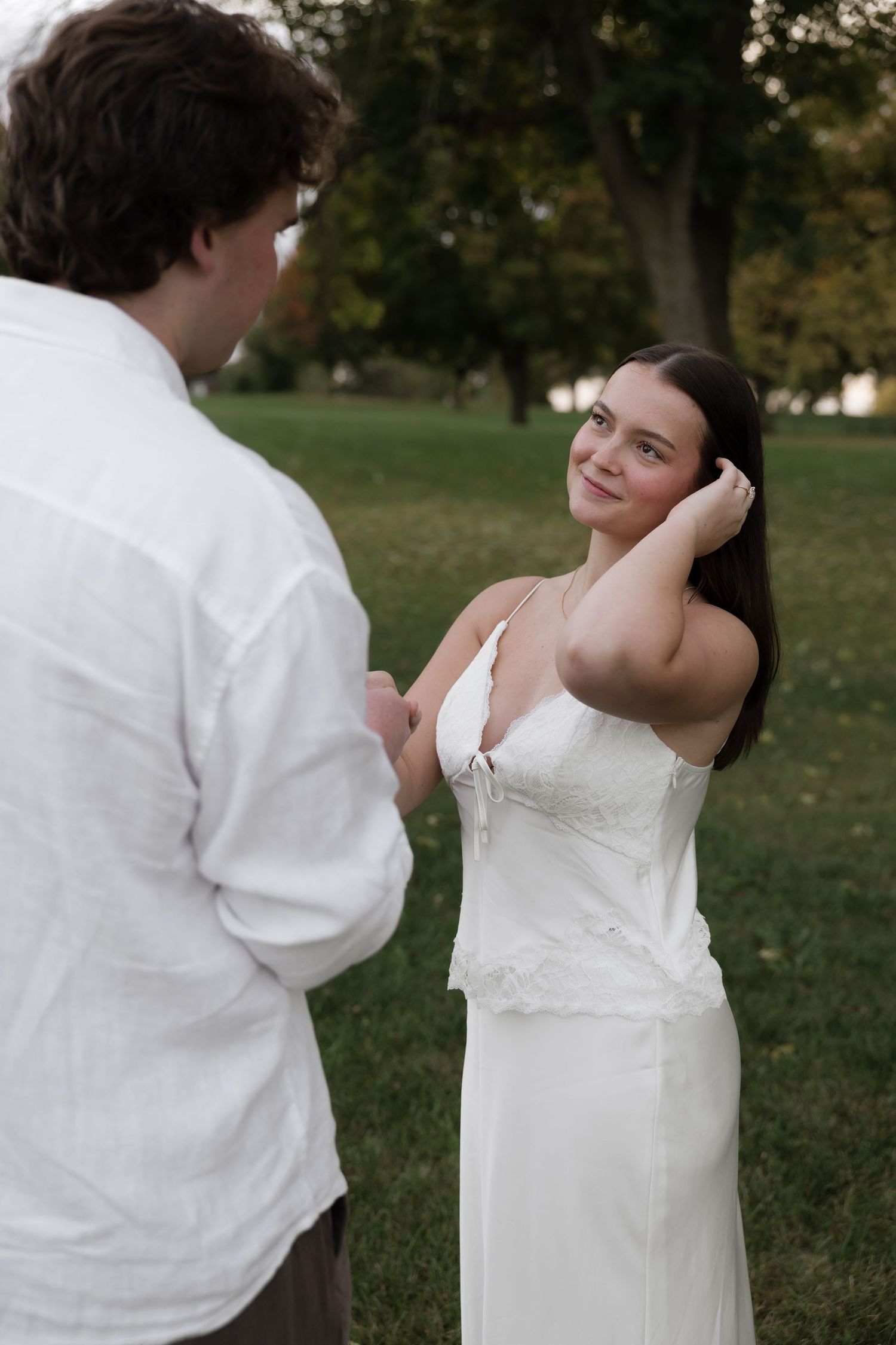 Woman in white lace dress smiling and tucking hair behind ear while holding hands with man in park.