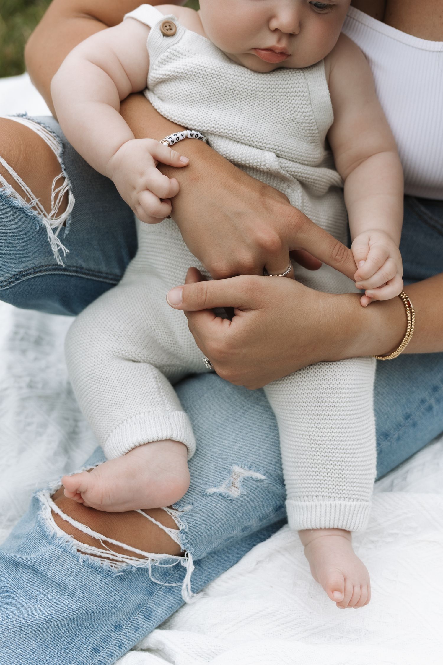 A baby in a knit romper sits on a parent's lap while another adult gently holds the infant's hands outdoors.
