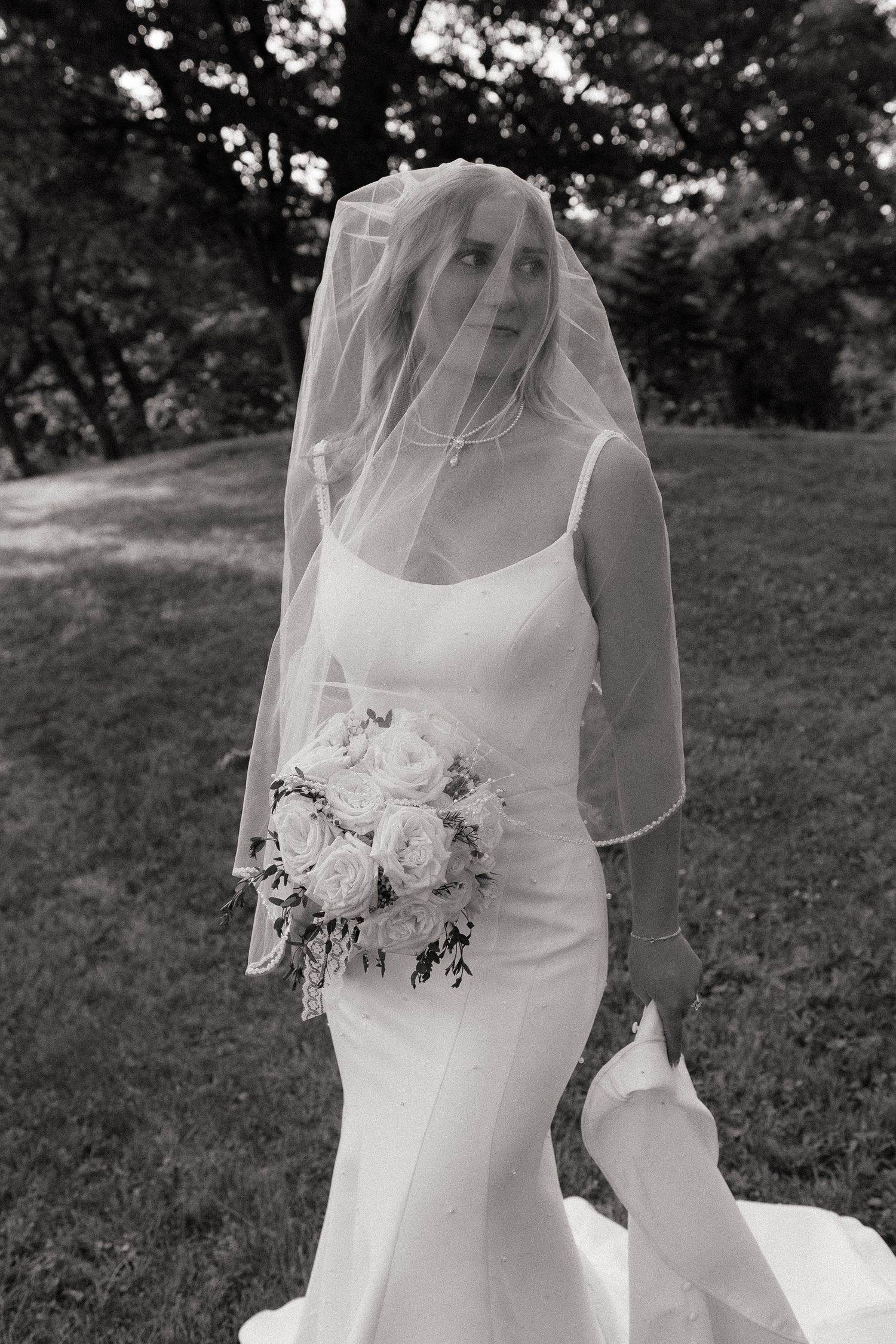 Black and white bridal portrait of bride in fitted gown with veil over face holding white rose bouquet outdoors.