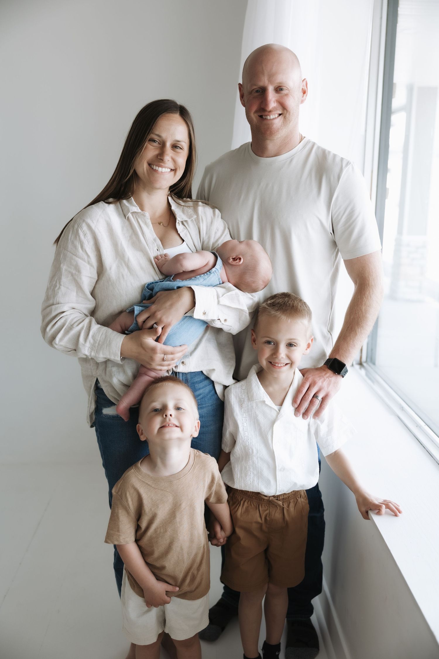 Happy family of five with newborn baby posing together by a bright window.